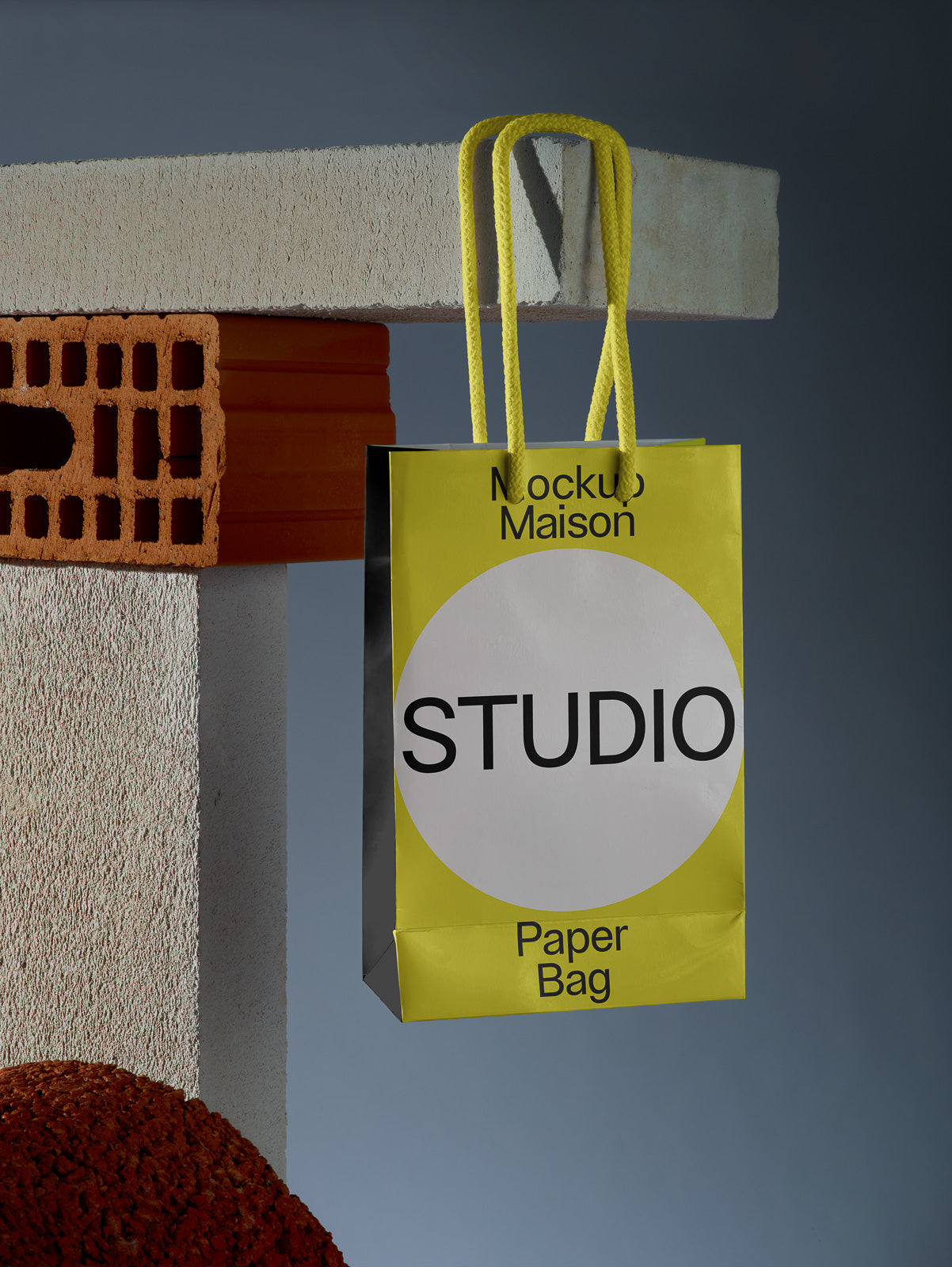 PSD mockup of a yellow paper bag with black text "Studio" hanging from concrete and brick blocks, featuring bold handles against a neutral background.