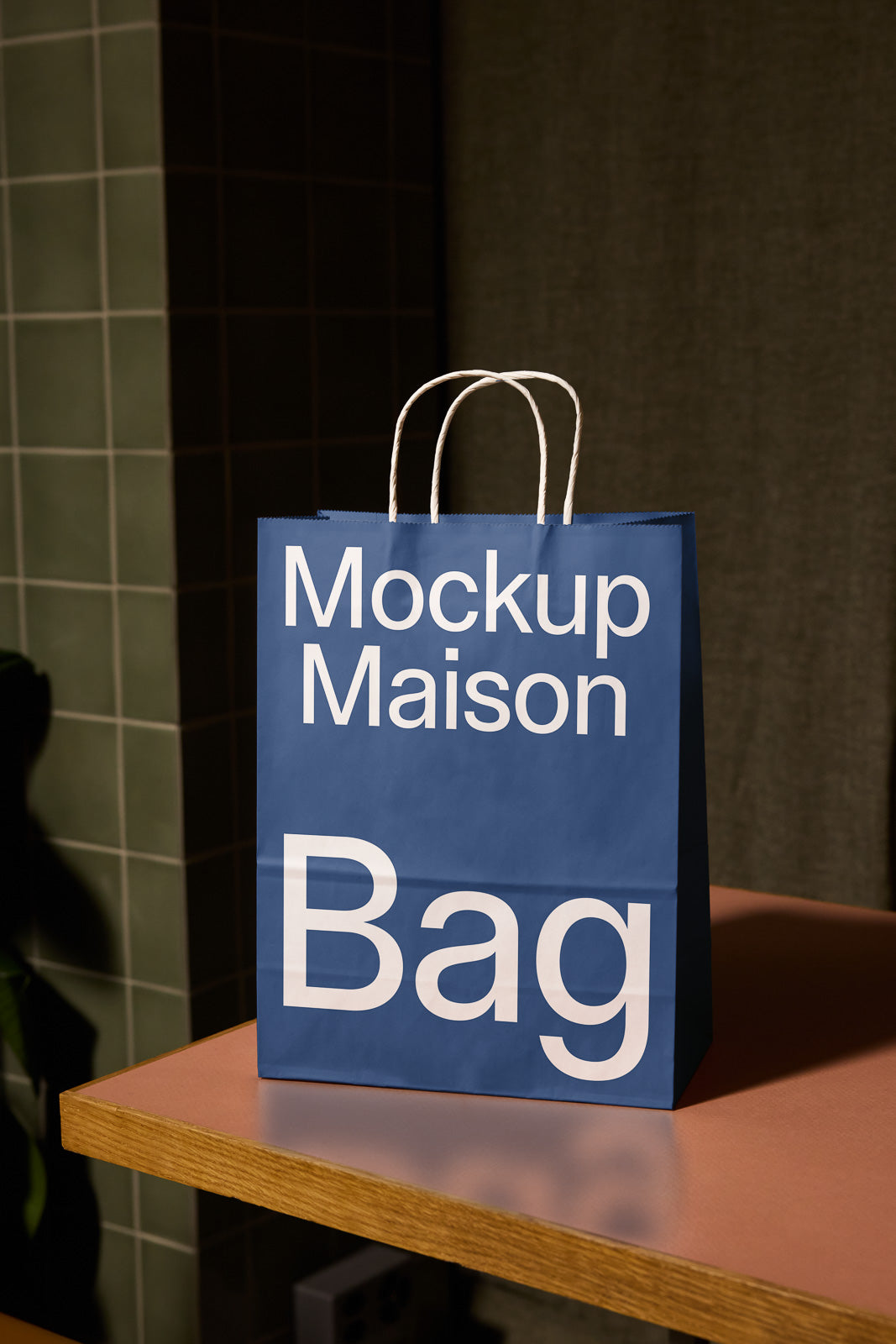 PSD mockup of a blue paper shopping bag with "Mockup Maison" in white text, positioned on a wooden table with a tile wall background.