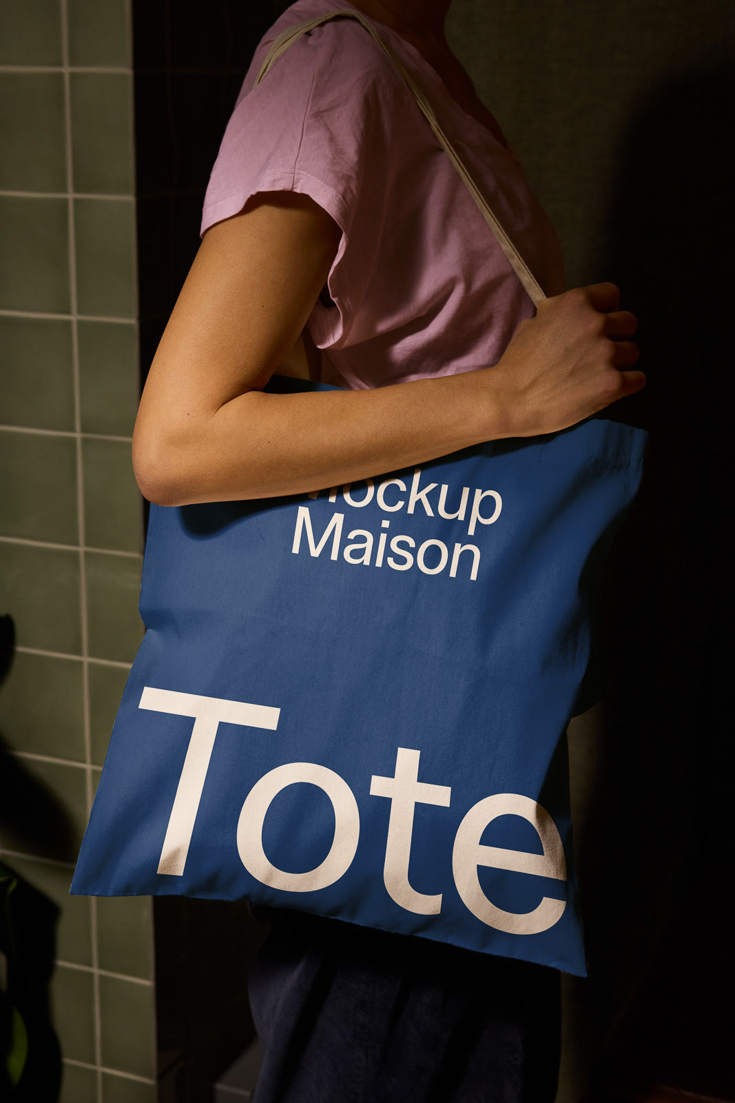 PSD mockup of a person holding a blue tote bag labeled "Mockup Maison" and "Tote" against a tiled background.