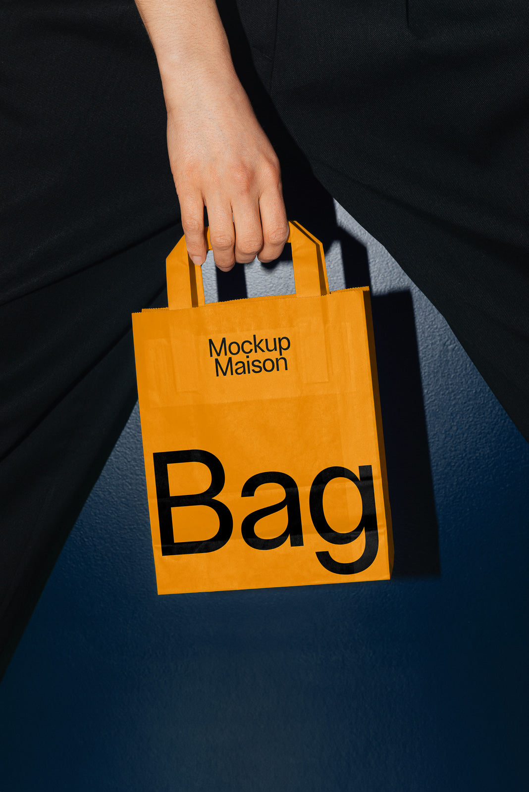 PSD mockup of a person holding an orange paper bag with "Mockup Maison" and "Bag" printed on it against a dark background.