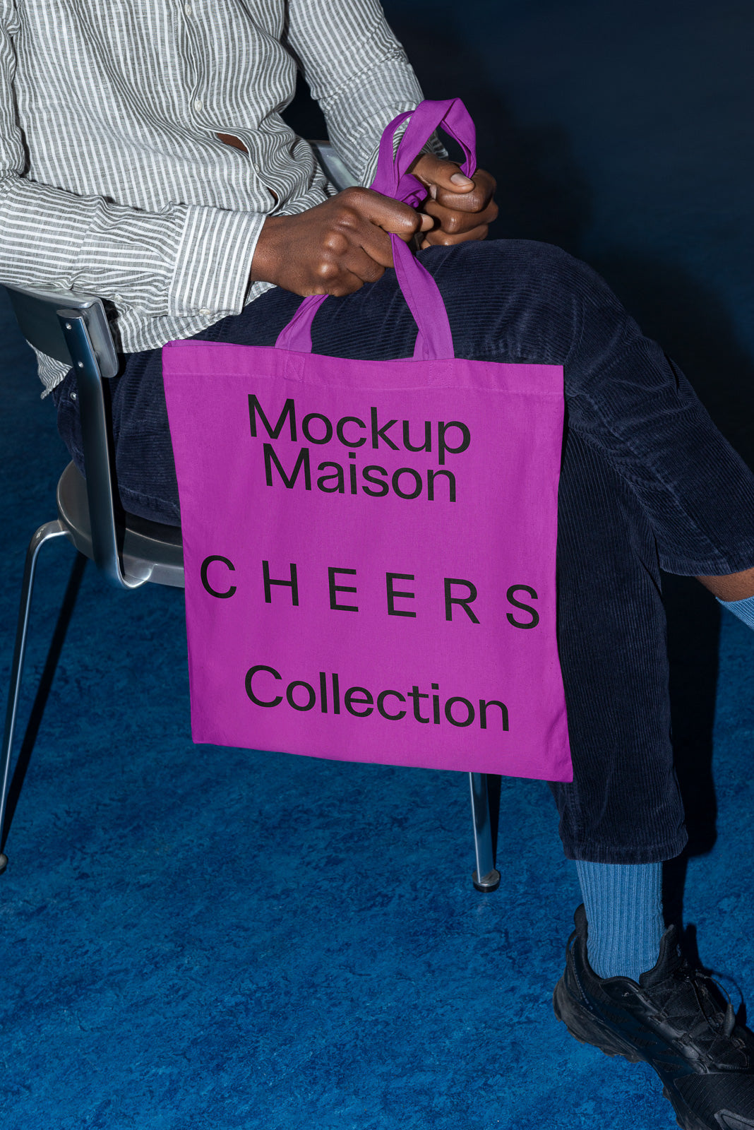 PSD mockup of a person holding a bright purple tote bag with black text reading "Mockup Maison CHEERS Collection," seated on a chair with blue floor beneath.