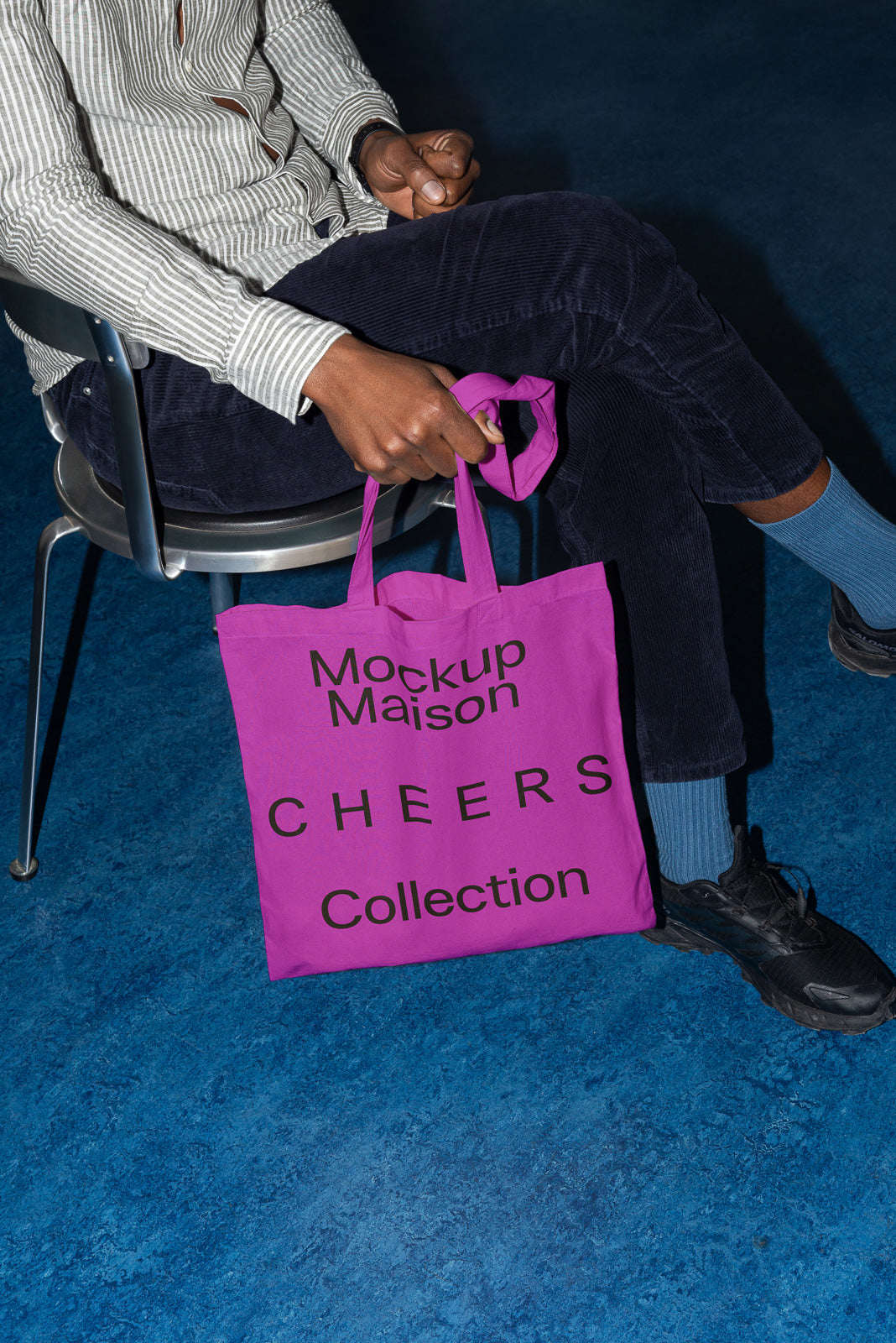 PSD mockup of a person sitting on a chair holding a magenta tote bag with "Mockup Maison Cheers Collection" printed on it, set against a blue floor.