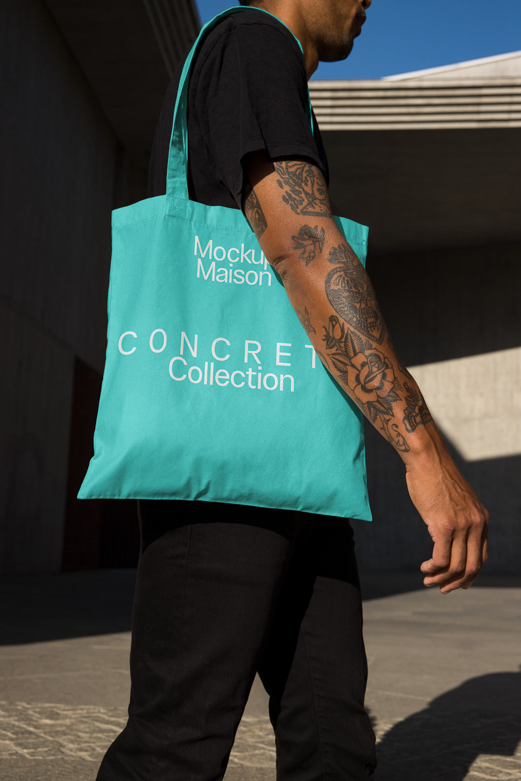 PSD mockup of a person holding a turquoise tote bag labeled "Mockup Maison CONCRETE Collection," against an outdoor urban backdrop.
