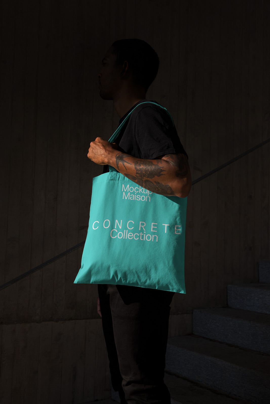 PSD mockup of a person holding a teal tote bag with "Mockup Maison Concrete Collection" printed on it, standing against a dark background.