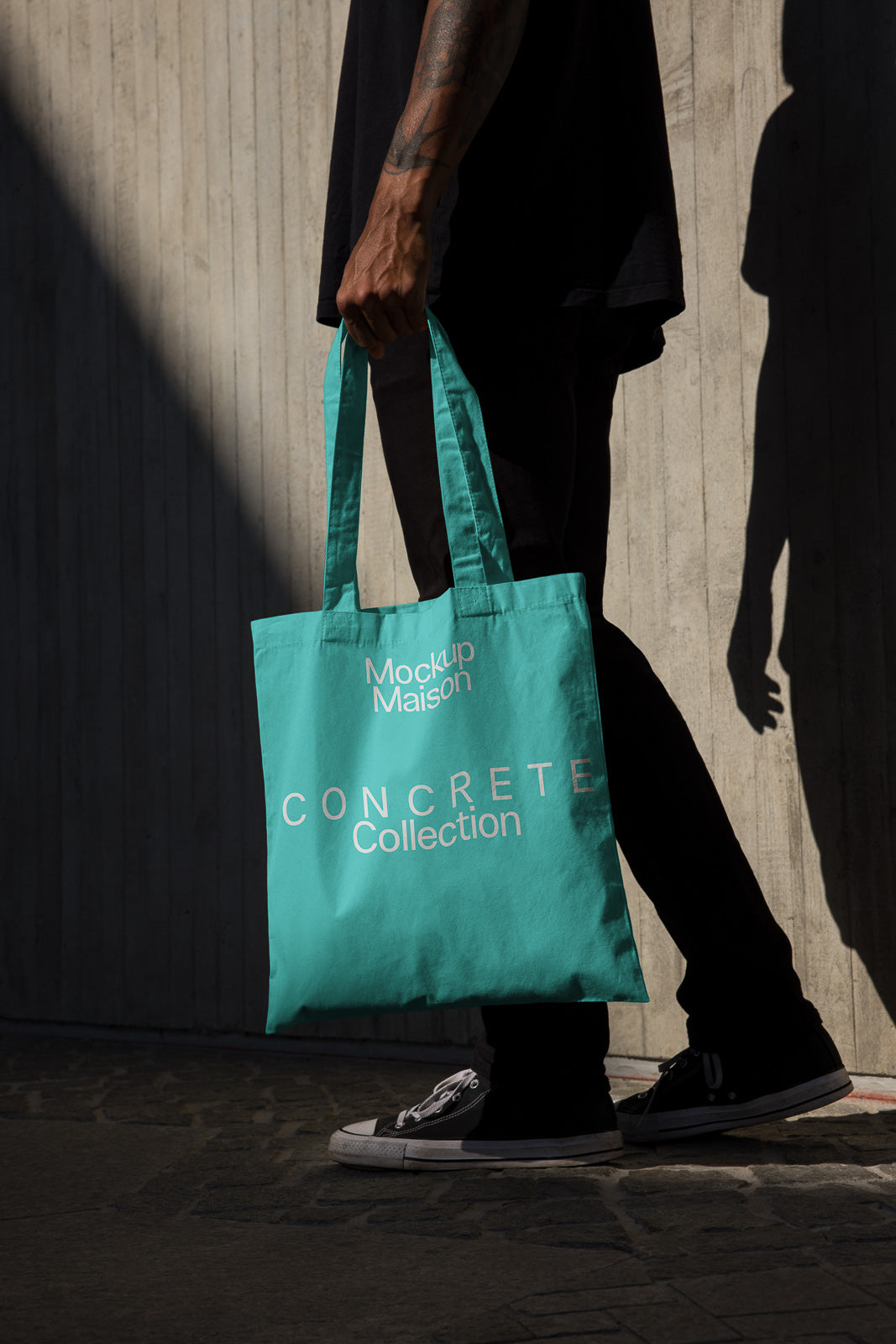 PSD mockup of a person holding a teal tote bag with "Concrete Collection" text, casting a shadow against a textured wall.