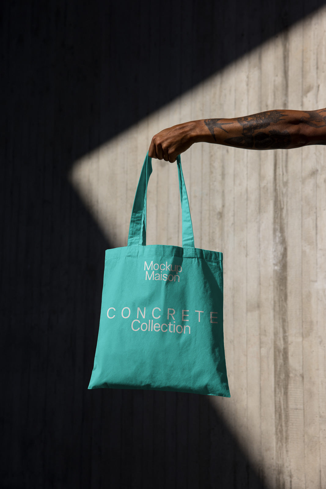 PSD mockup of a turquoise tote bag with "Mockup Maison" and "Concrete Collection" text, held by a tattooed arm against a textured concrete wall.