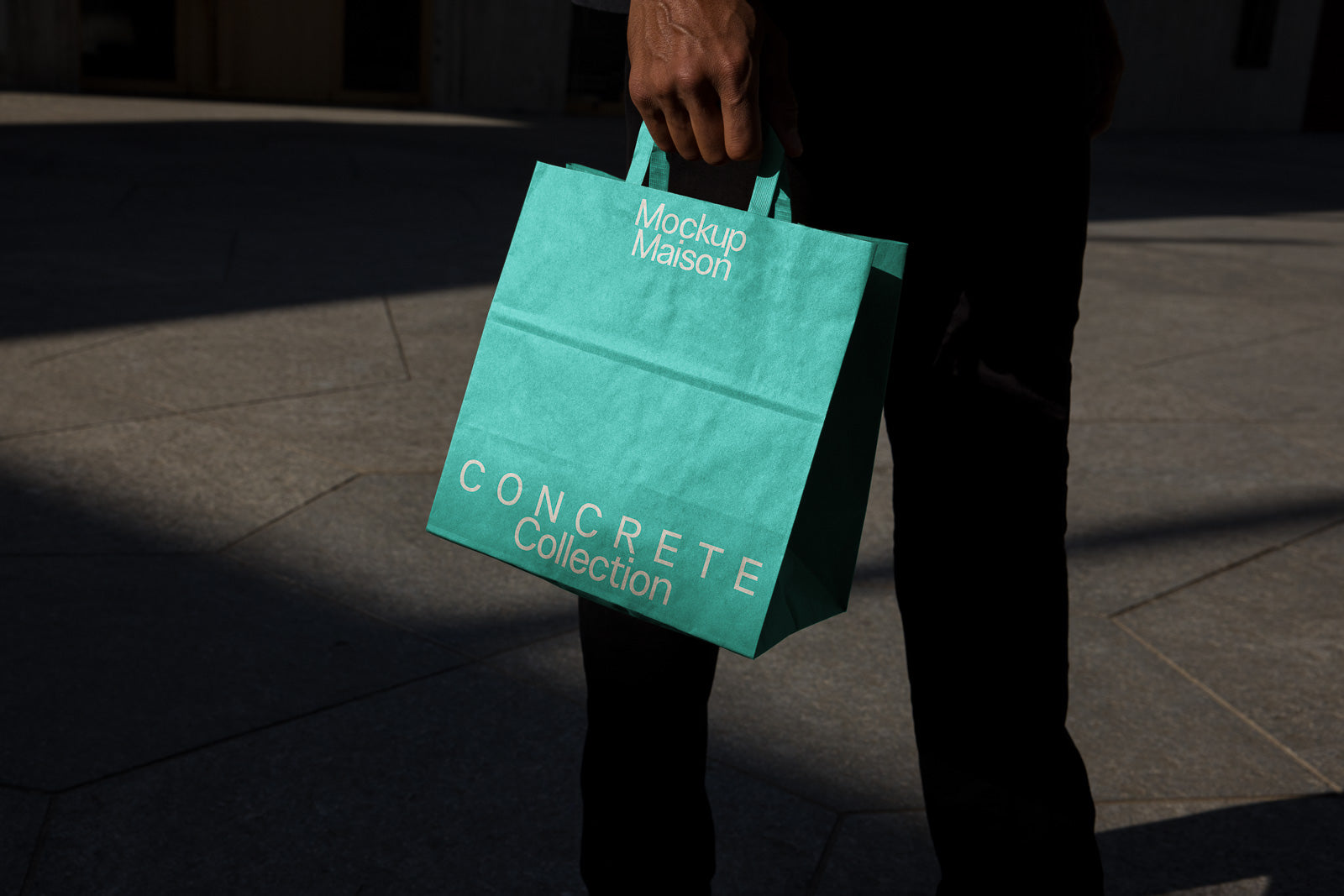 PSD mockup of a teal shopping bag with "Mockup Maison" and "Concrete Collection" text, held by a person against a stone pavement.