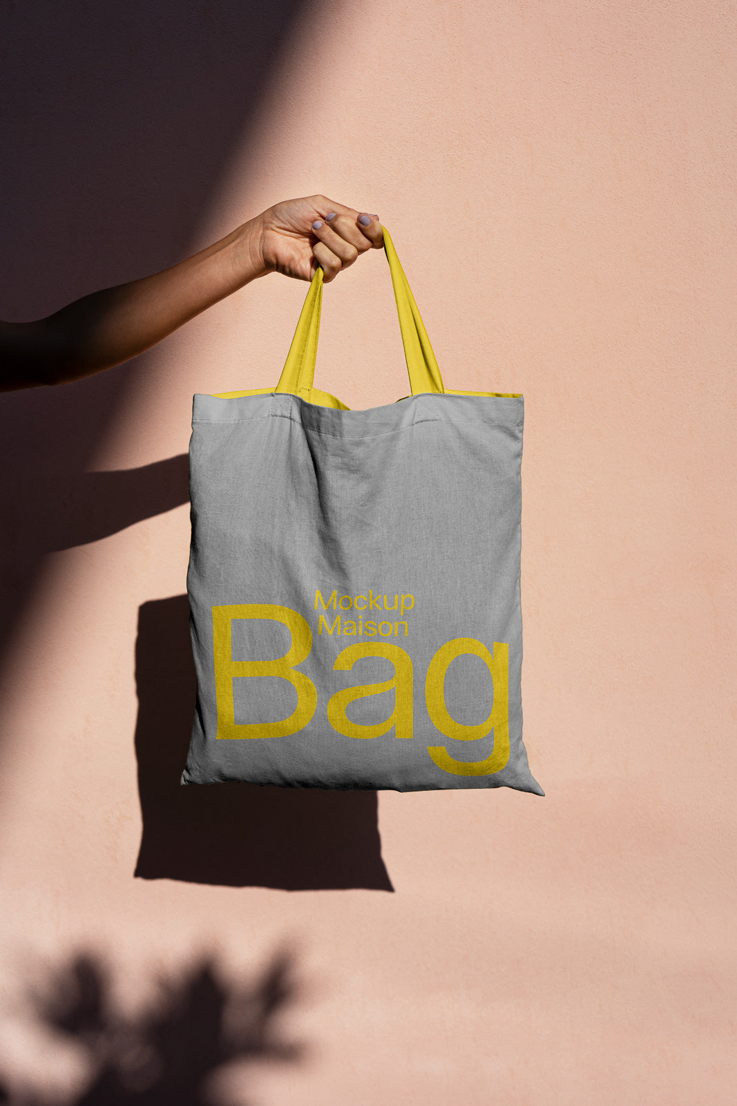 PSD mockup of a hand holding a gray tote bag with yellow handles against a peach wall. The bag features yellow text "Mockup Maison Bag."