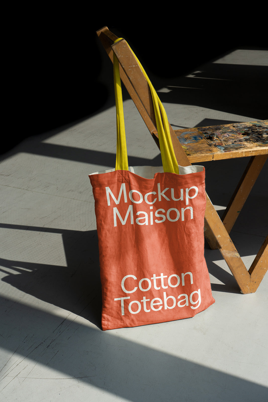 PSD mockup of a red cotton tote bag with white text leaning against a wooden chair in a sunlit room with a concrete floor.