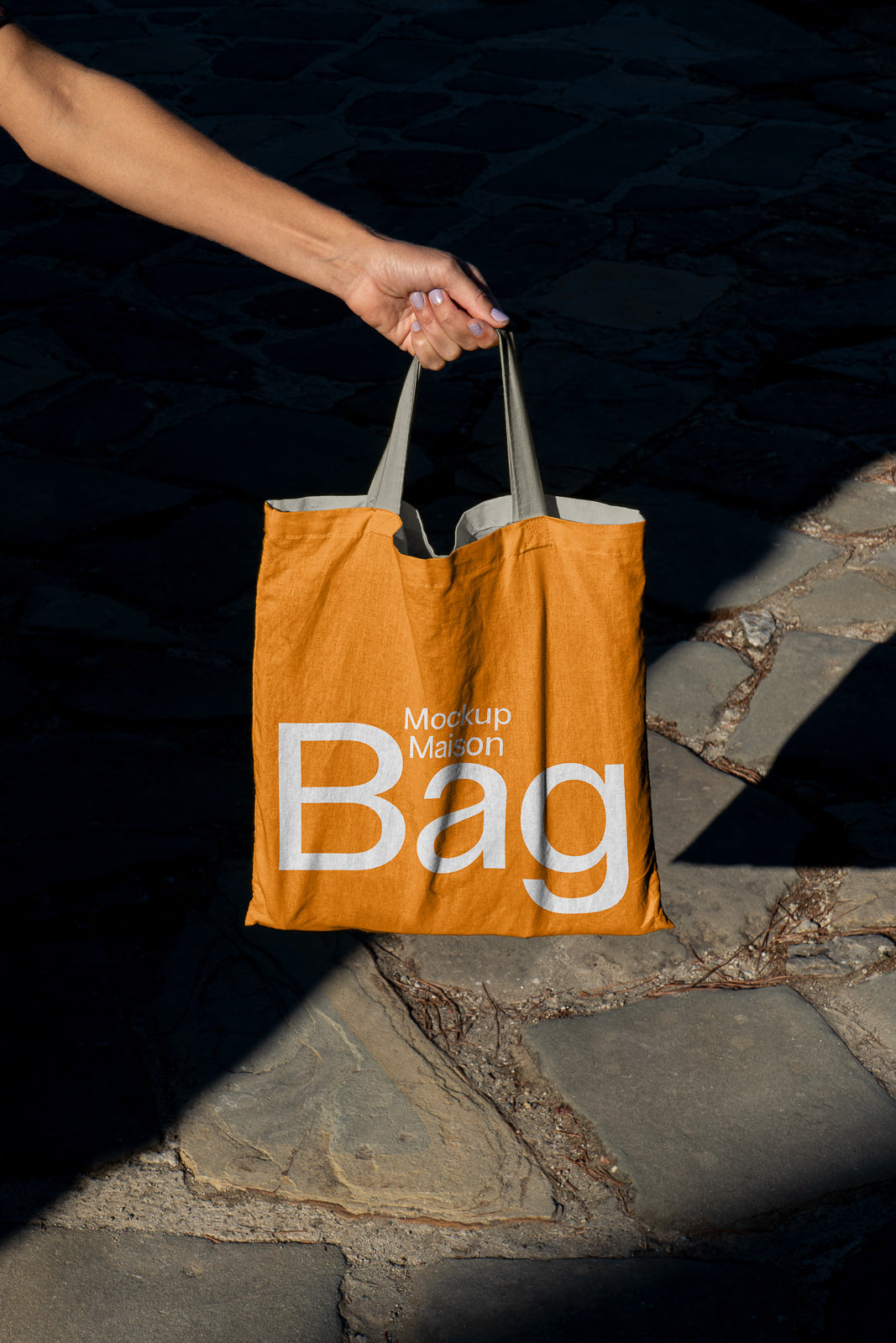 PSD mockup of a person holding an orange tote bag with "Mockup Maison Bag" text, casting a shadow on a stone pavement.