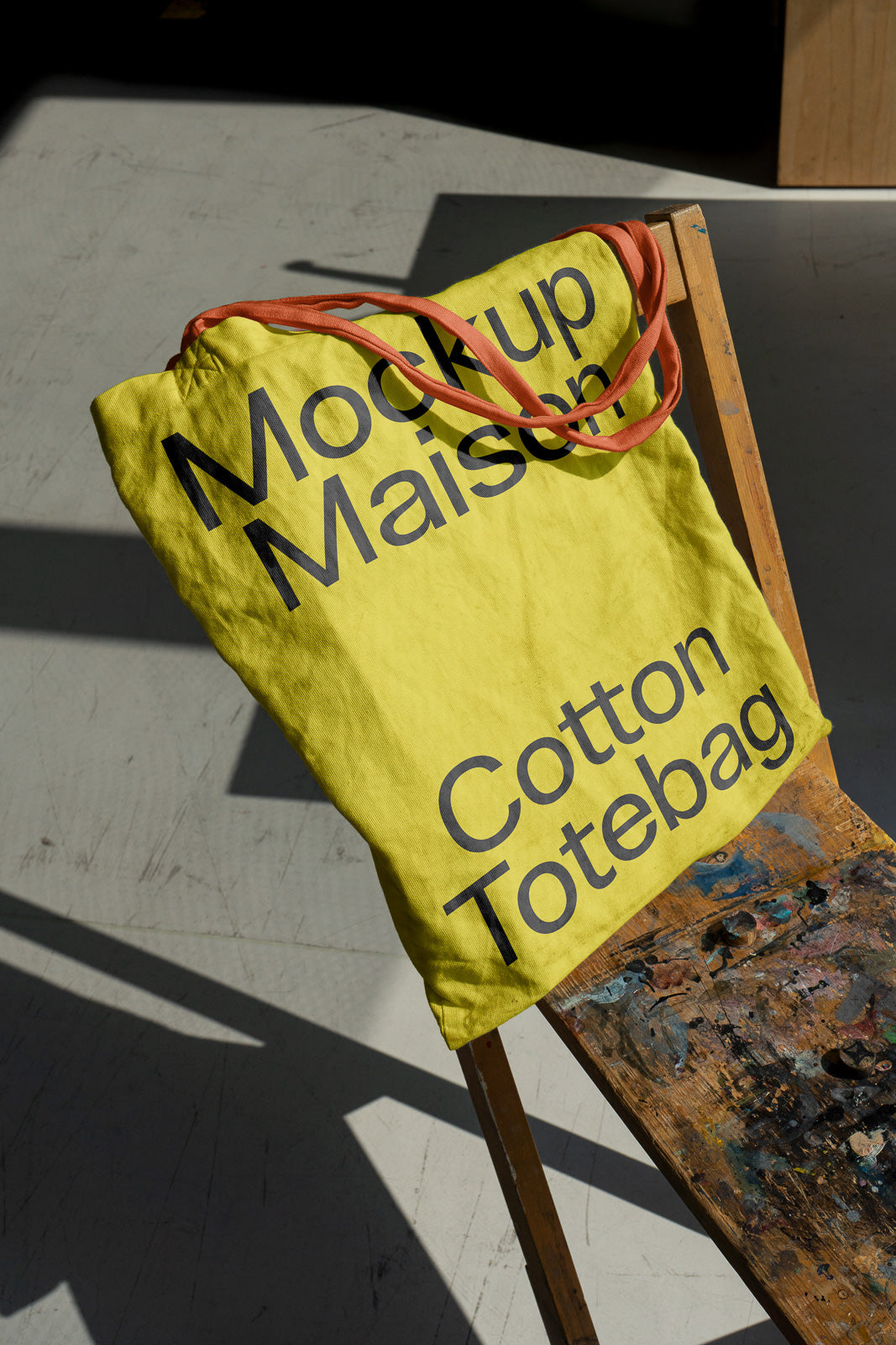PSD mockup of a yellow cotton tote bag with the text "Mockup Maison" draped over a wooden chair, casting shadows on a sunlit floor.
