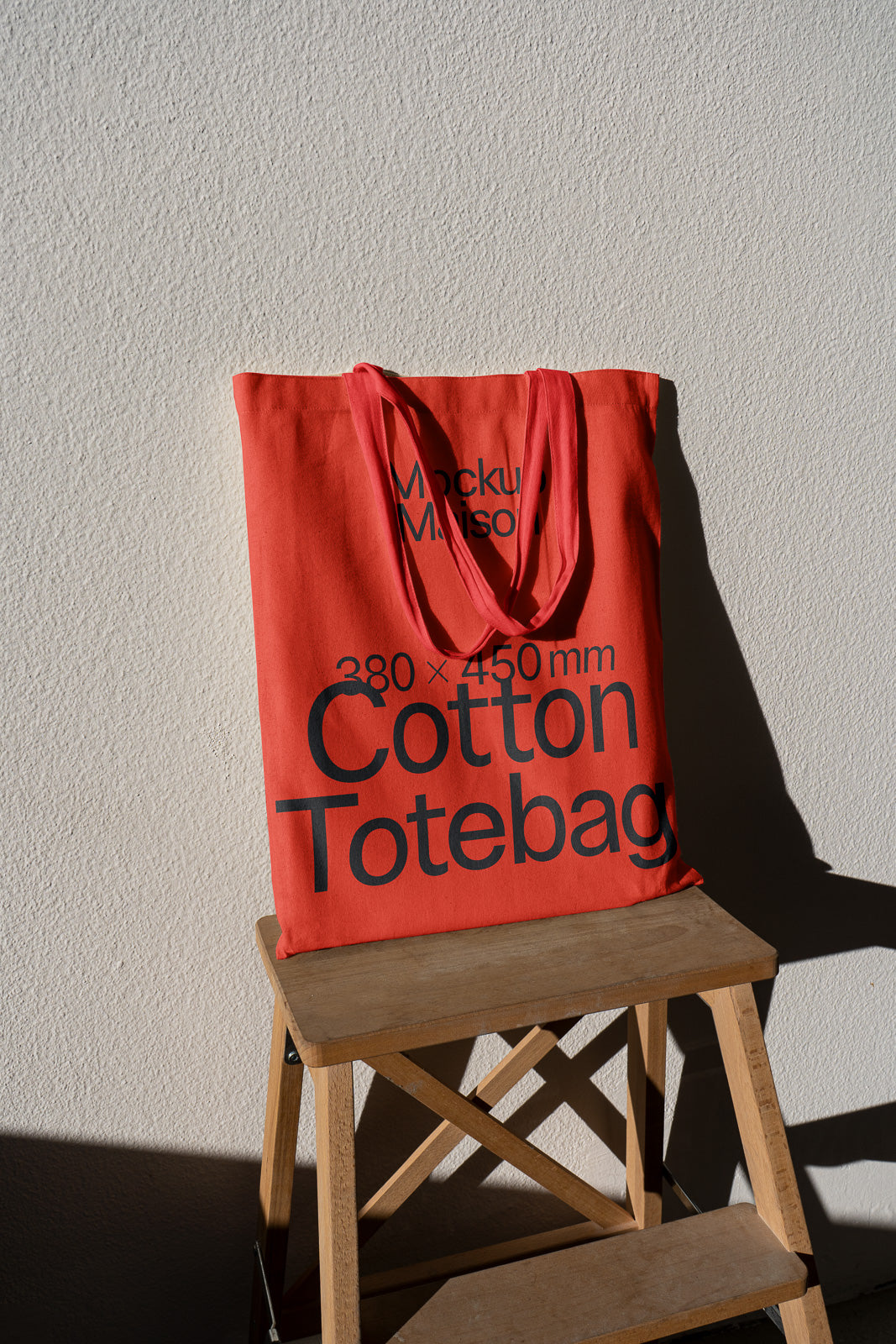 PSD mockup of a red cotton tote bag with dimensions 380 x 450mm printed on it, hanging on a wooden chair against a textured wall.