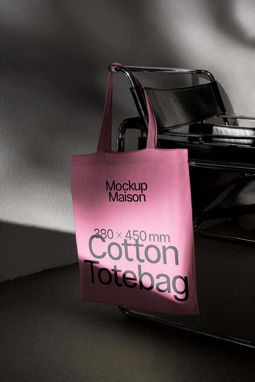 PSD mockup of a pink cotton tote bag with text "Mockup Maison 380 x 450 mm" hanging on a modern black chair against a textured wall.