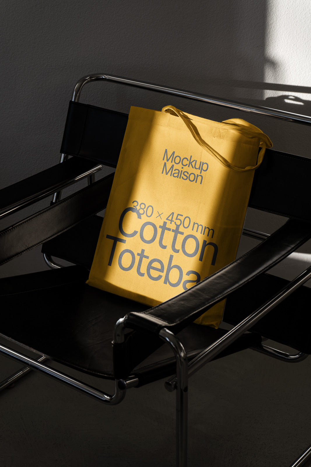 PSD mockup of a yellow cotton tote bag with "Mockup Maison, 380 x 450 mm" printed, hanging on a black leather chair in soft lighting.