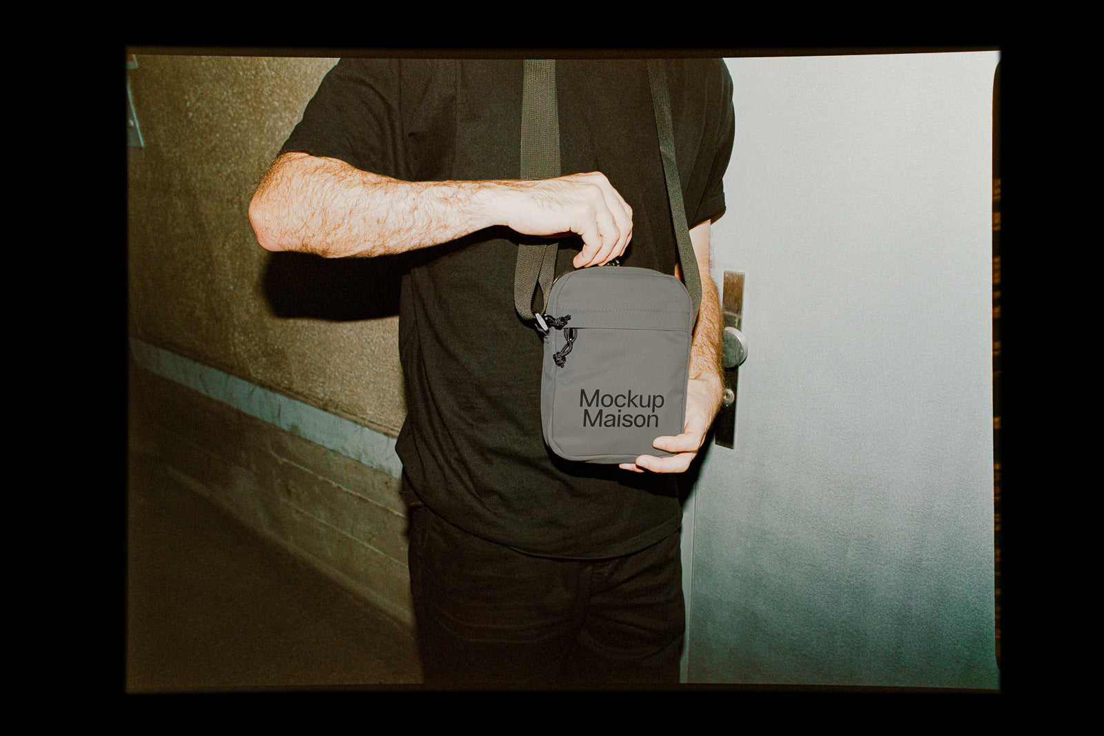 PSD mockup of a person holding a small gray crossbody bag with "Mockup Maison" text printed on it, standing near a door.