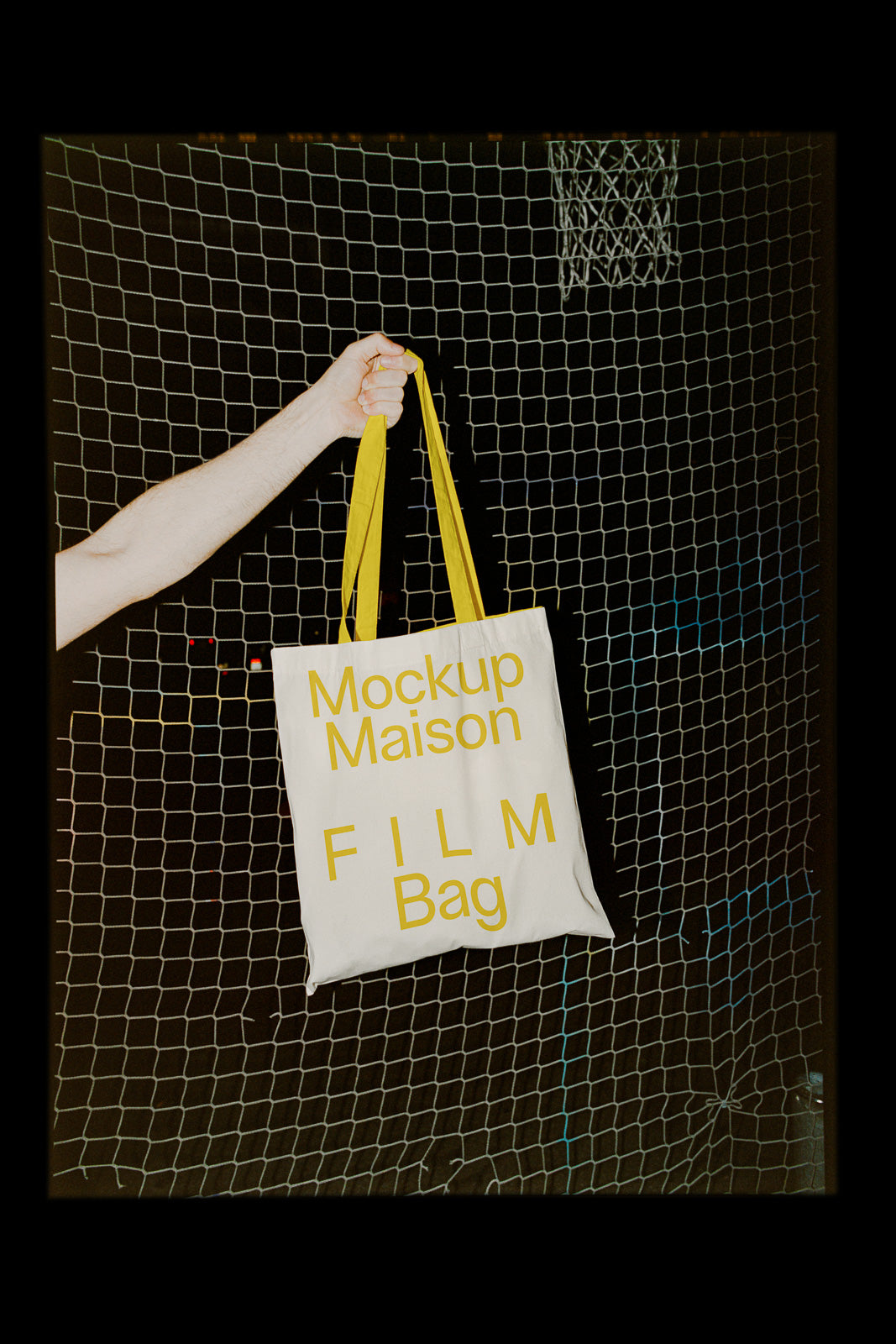 PSD mockup of a tote bag with yellow text “Mockup Maison FILM Bag” held by a hand against a netted background.
