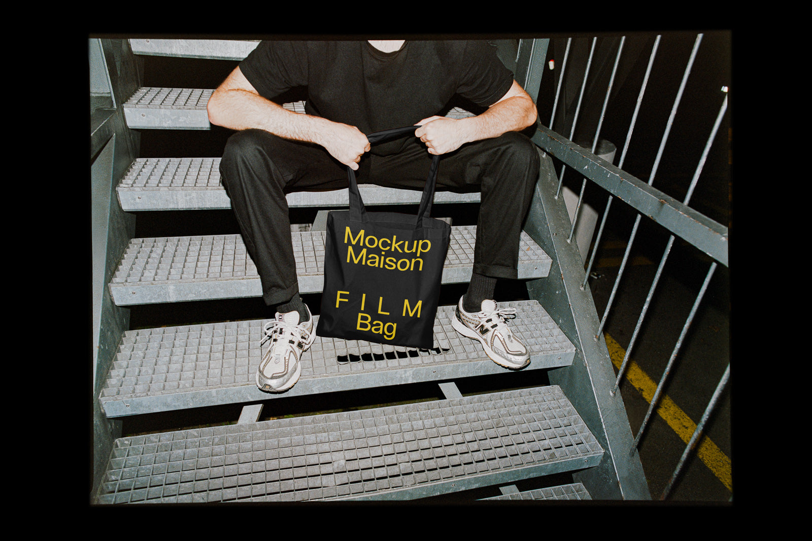 PSD mockup of a person sitting on metal stairs holding a black tote bag with yellow text reading "Mockup Maison FILM Bag."