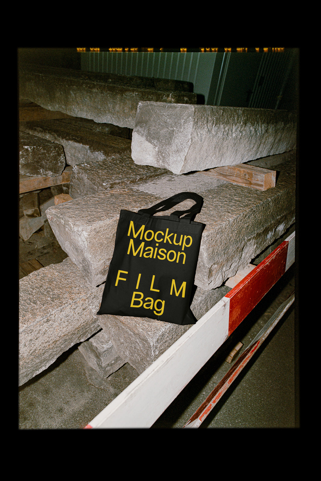 PSD mockup of a black tote bag with bold yellow text "Mockup Maison FILM Bag" placed on large stone blocks in an industrial setting.