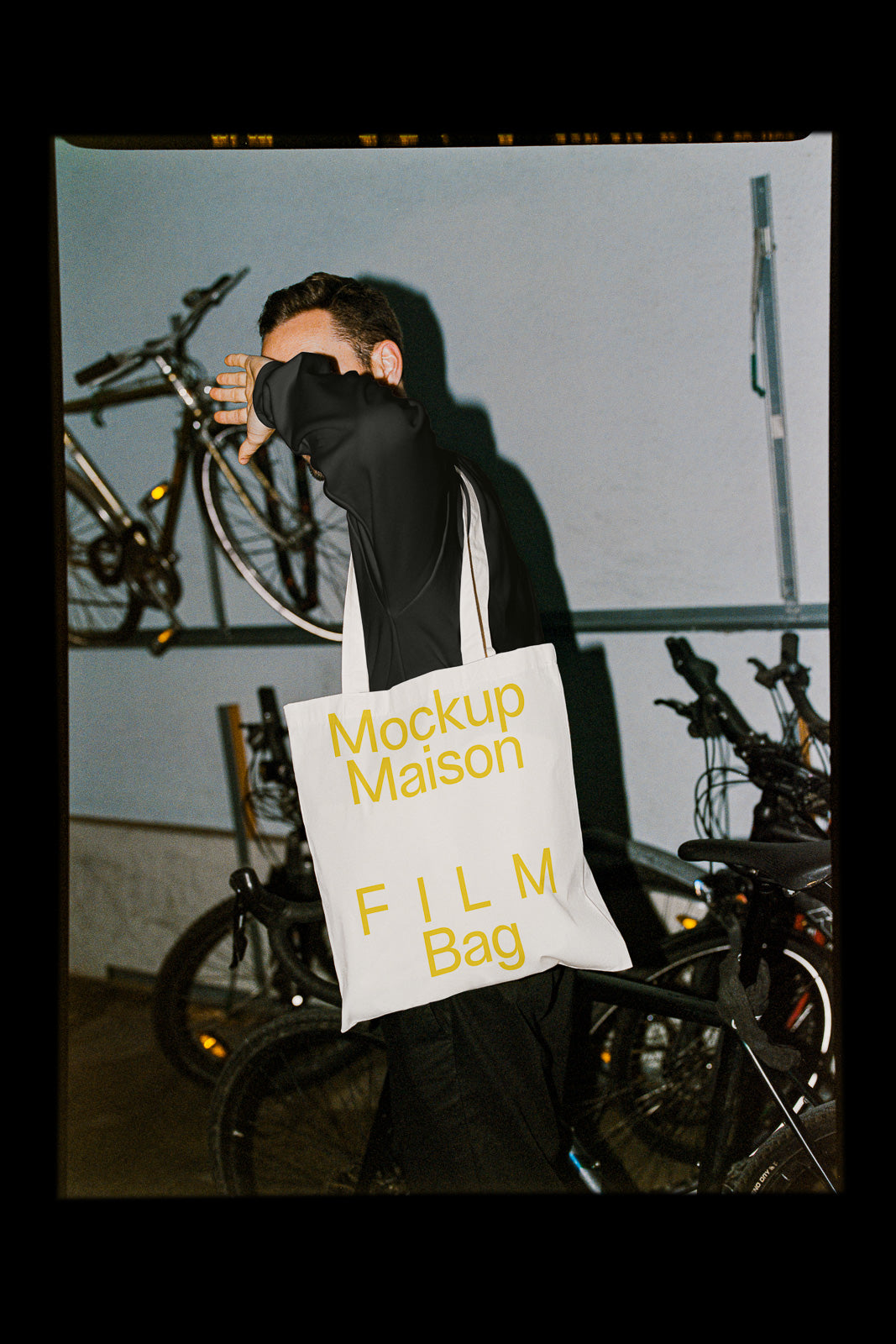 PSD mockup of a tote bag with yellow text "Mockup Maison FILM Bag" carried by a person standing near bicycles indoors.