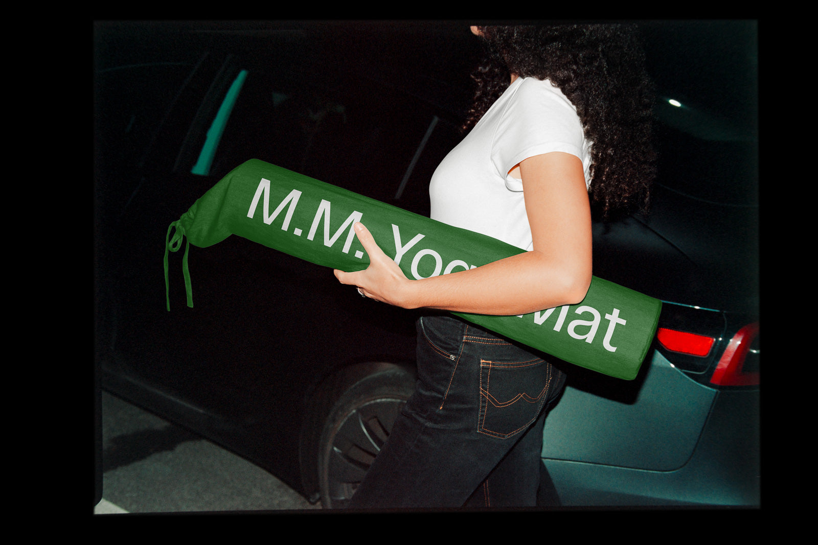 PSD mockup of a person holding a green bag with "M.M. Yoga Mat" printed on it, standing near a car at night.