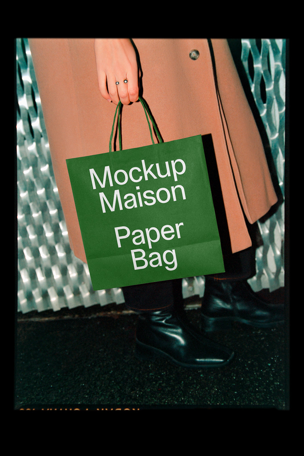 PSD mockup of a person holding a green paper bag labeled "Mockup Maison Paper Bag" against a textured metallic background, wearing a coat and black boots.