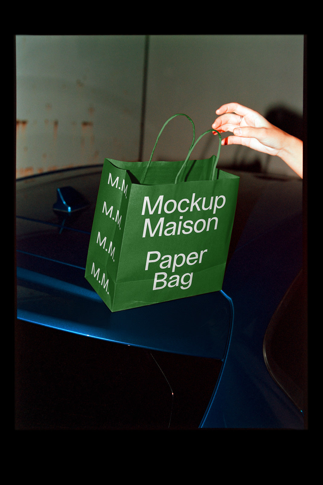 PSD mockup of a green paper bag with "Mockup Maison" text, resting on a blue car with a hand reaching to grab it, creating a dynamic scene.
