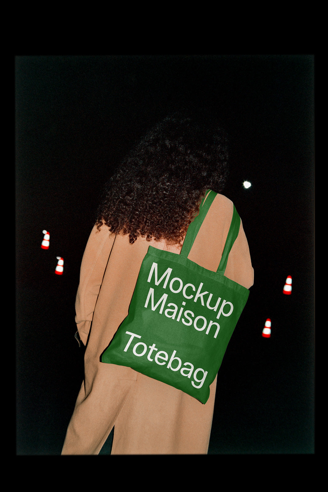 PSD mockup of a green tote bag labeled "Mockup Maison Totebag," carried by a person in a tan coat with a dark background featuring traffic cones.