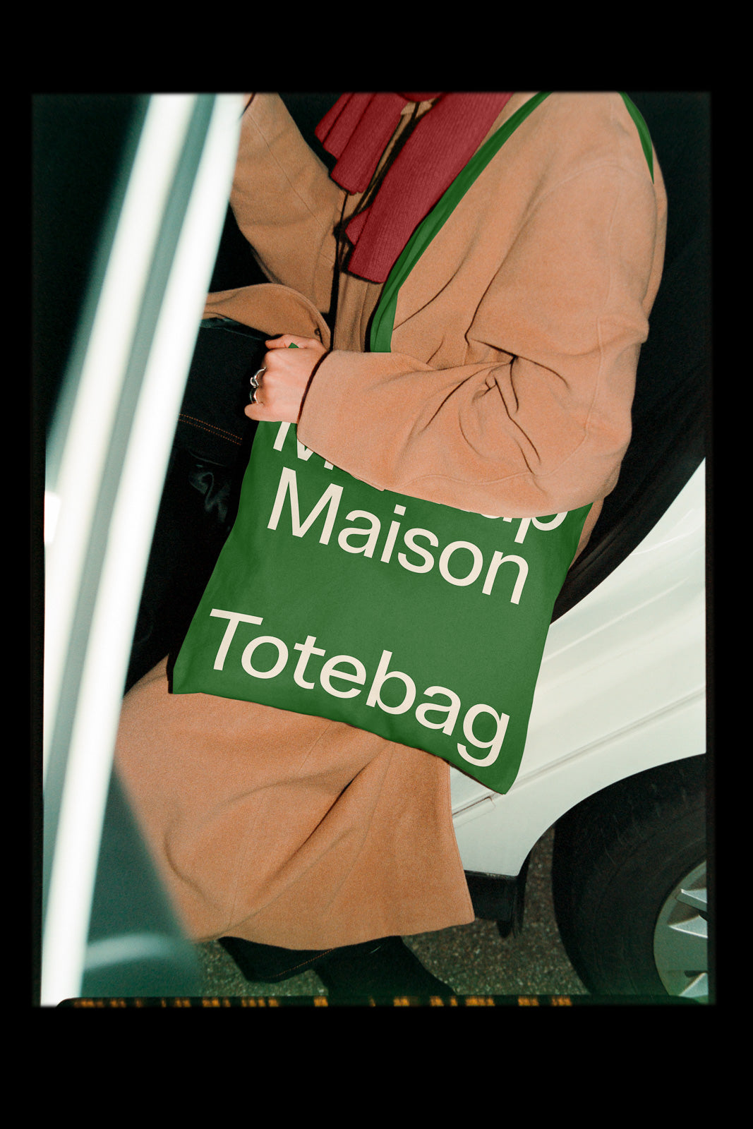 PSD mockup of a green tote bag with bold white text, held by a person wearing a tan coat and red scarf, positioned beside a car door.