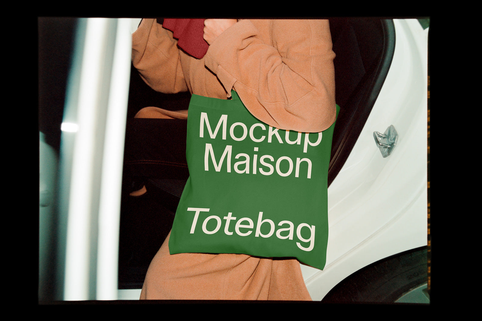 PSD mockup of a green tote bag with the text “Mockup Maison Totebag” being held by a person in a tan coat next to a white car.