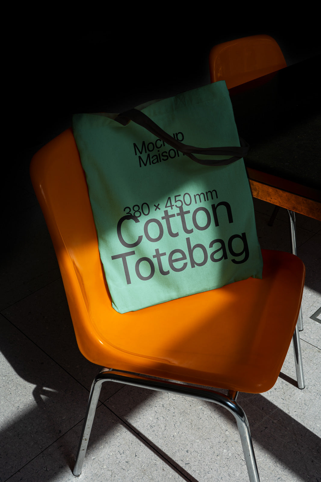 PSD mockup of a green cotton tote bag with text "380 x 450 mm" on an orange chair in a dimly lit setting.