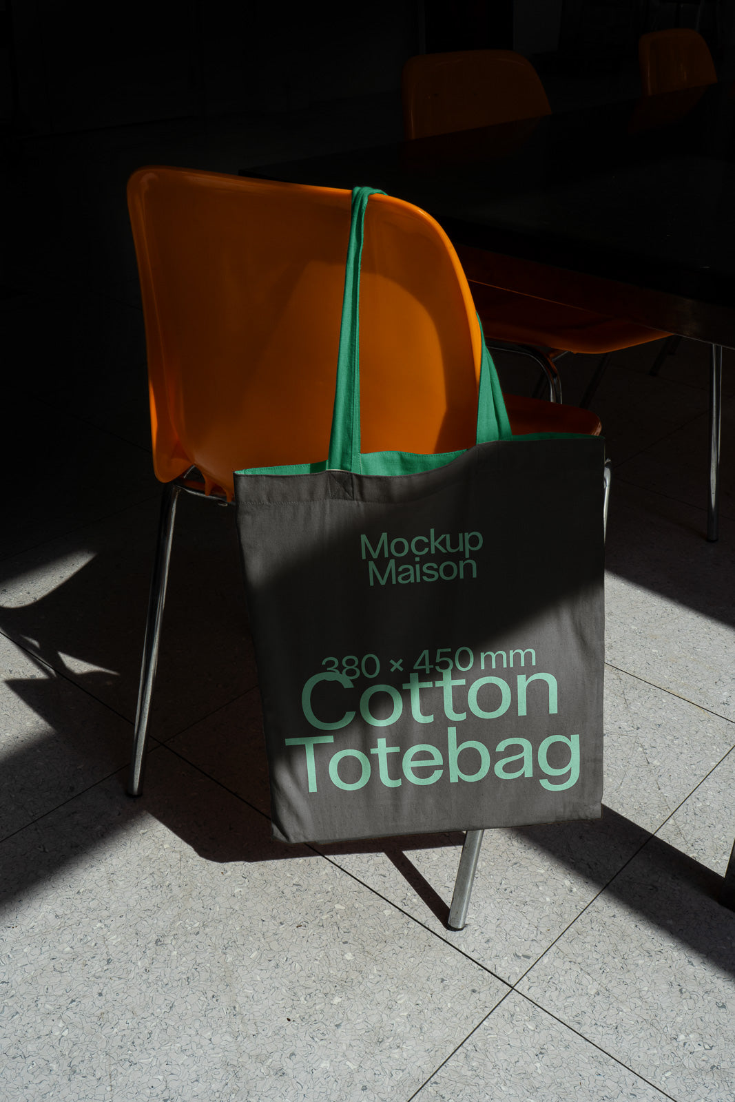 PSD mockup of a cotton tote bag with green handles and text displaying dimensions, hanging on an orange chair, casting shadows on a tiled floor.