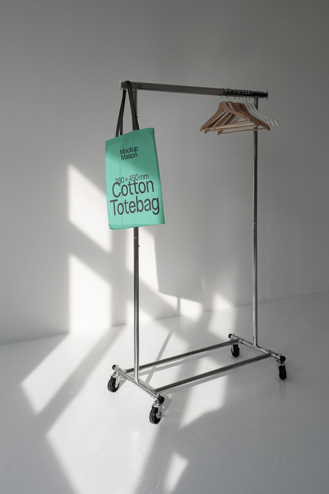PSD mockup of a cotton tote bag hanging from a garment rack with wooden hangers, casting shadows on a white floor and wall.