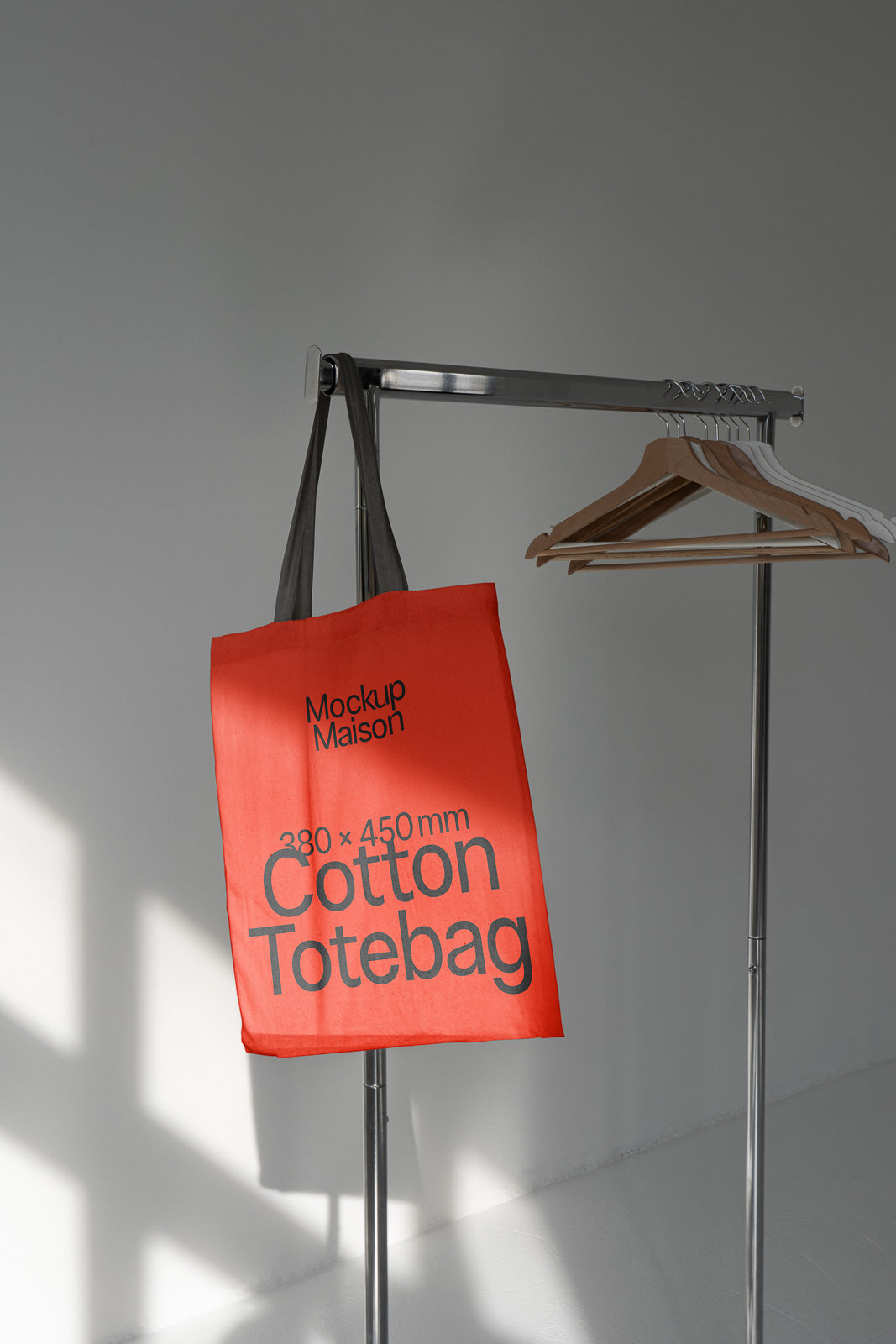 PSD mockup of a cotton tote bag with the text "Mockup Maison 380 x 450 mm Cotton Totebag," hanging on a metal rack with wooden hangers.
