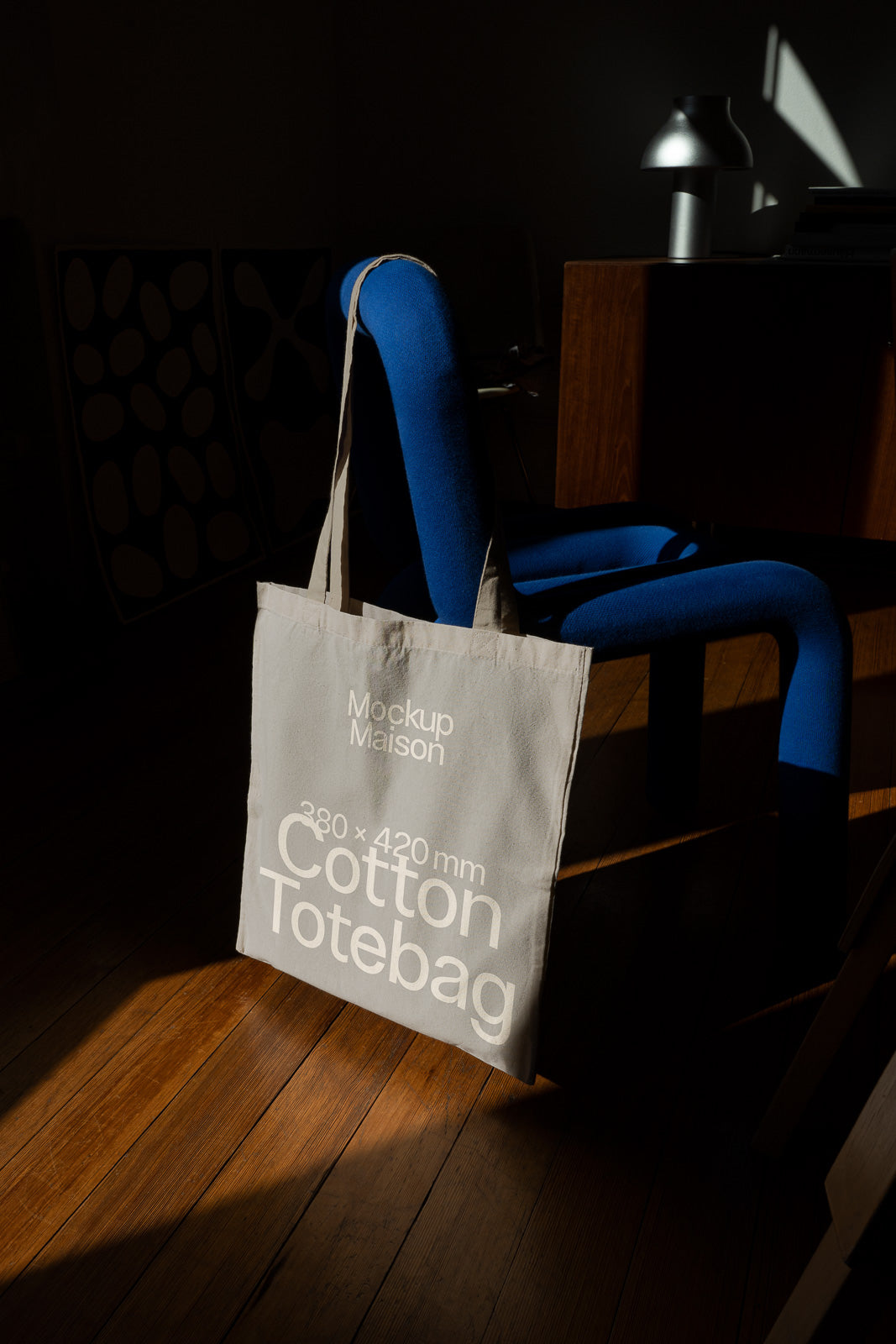 PSD mockup of a cotton tote bag hanging on a blue chair in a room with wooden flooring, featuring the dimensions 380 x 420 mm.