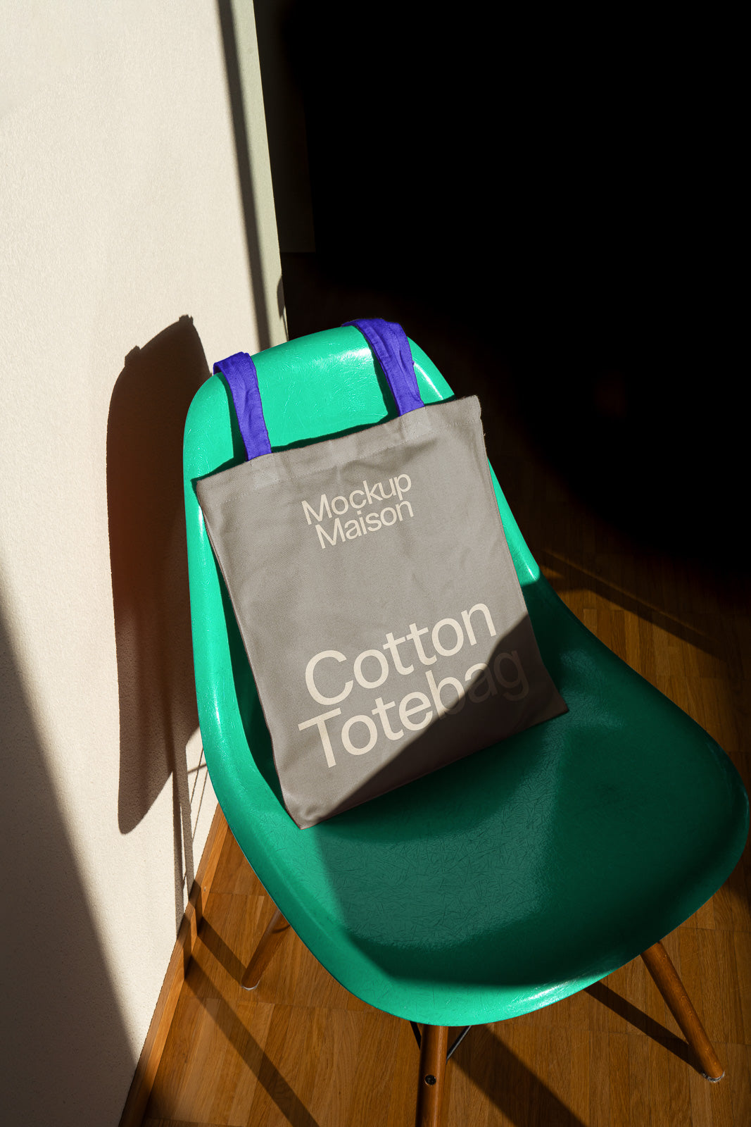 PSD mockup of a cotton tote bag with "Mockup Maison" text, placed on a green chair with blue handles in a room with wooden flooring.
