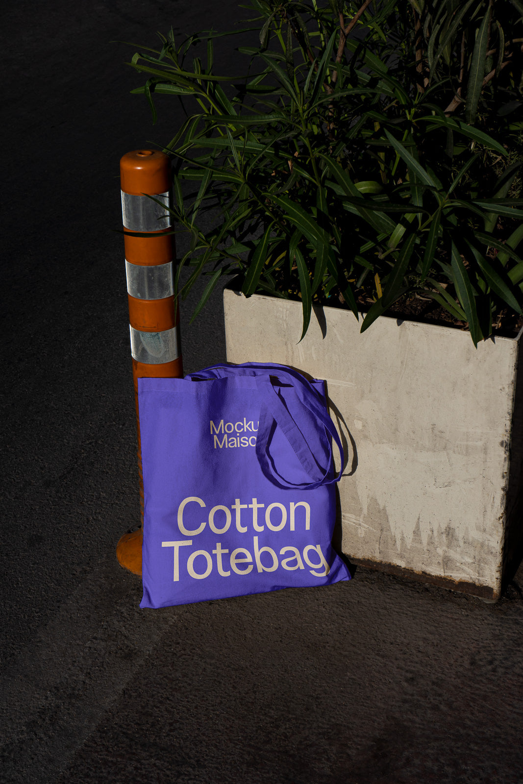 PSD mockup of a purple cotton tote bag leaning against a concrete planter and traffic post, showcasing minimalist text design.