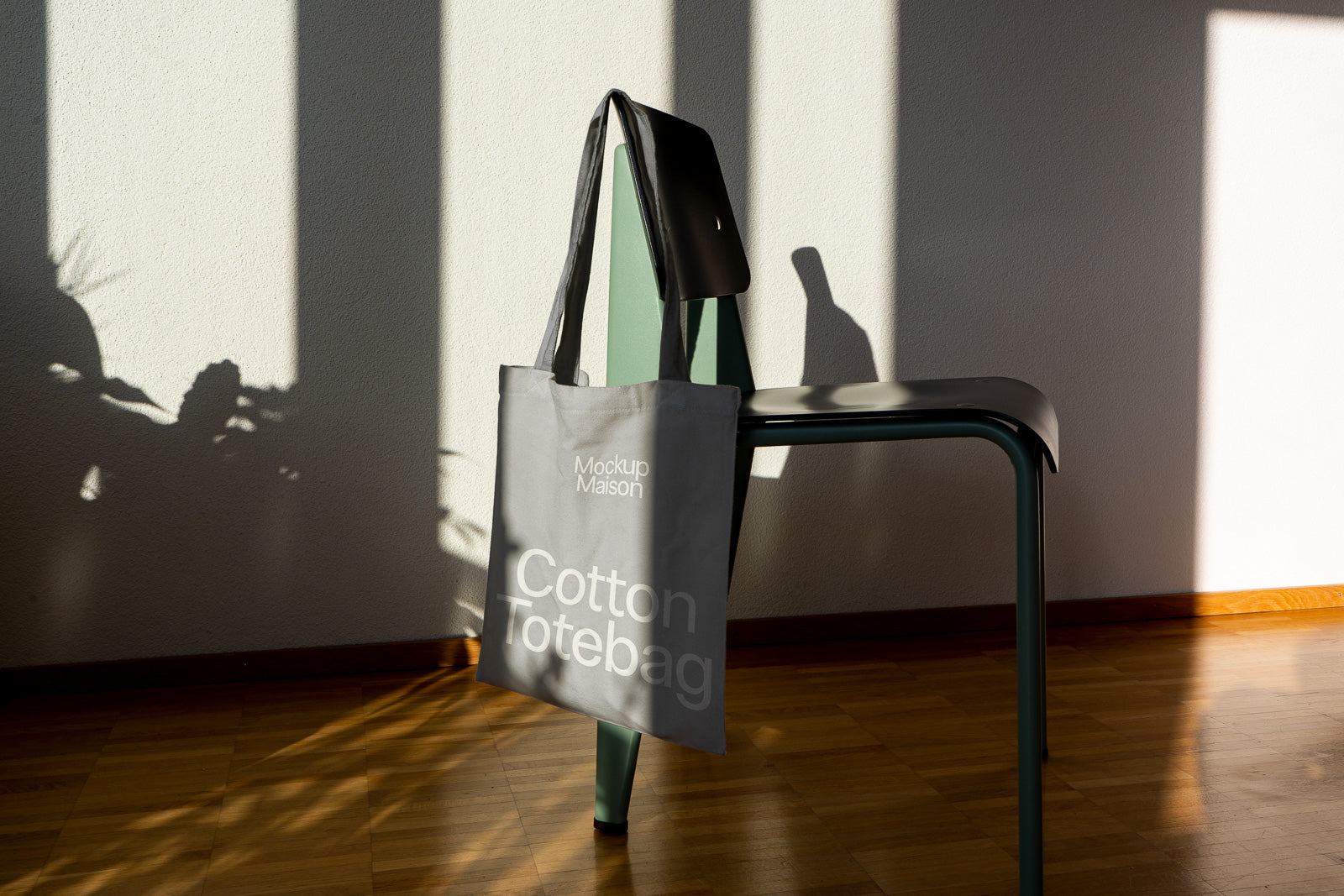 PSD mockup of a gray cotton tote bag hanging on a green chair against a sunlit wall, casting distinct shadows on the wooden floor.