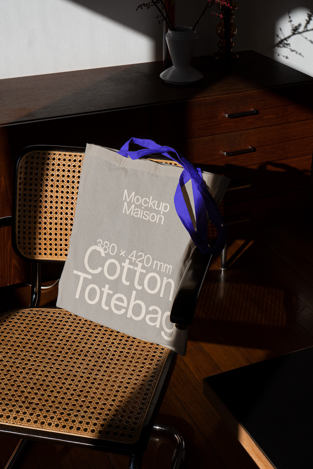 PSD mockup of a cotton tote bag with "Mockup Maison" branding, resting on a wooden chair with a woven seat, highlighting its texture and size.