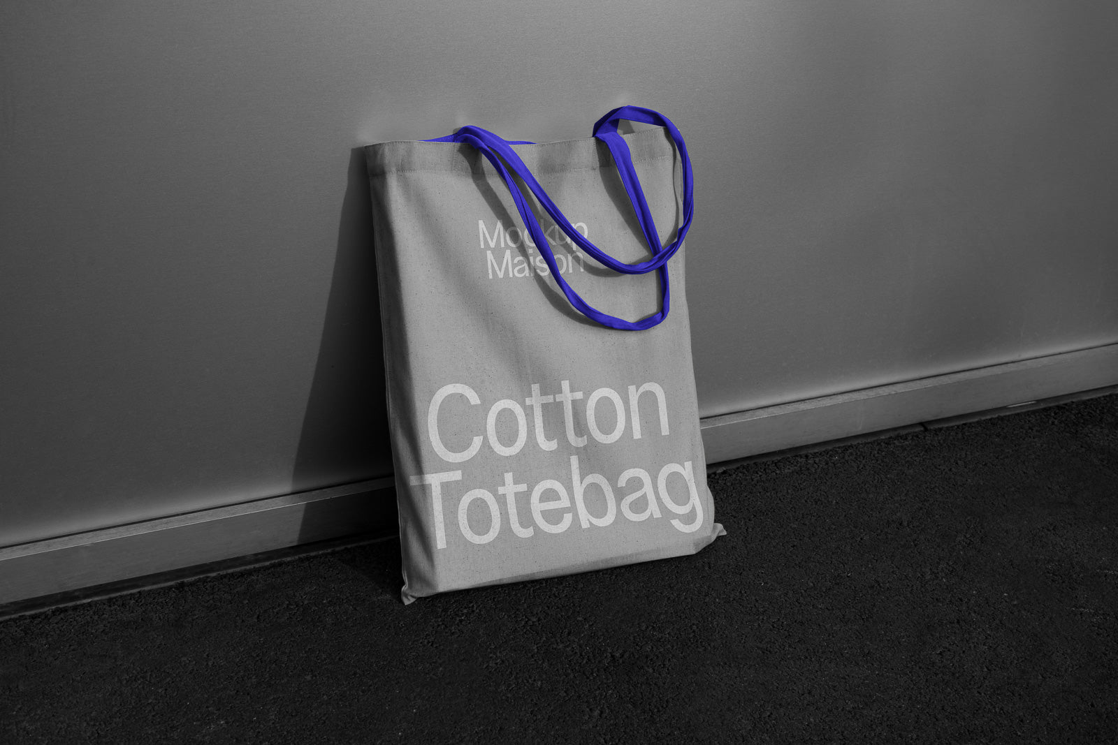 PSD mockup of a gray cotton tote bag with bold white text, featuring blue handles, leaning against a gray wall on a dark floor.