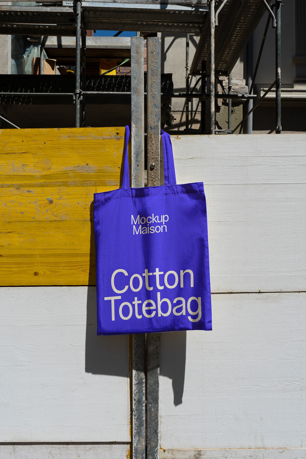PSD mockup of a bright purple cotton tote bag hanging on a textured metal pole against a painted wooden wall with a shadow.