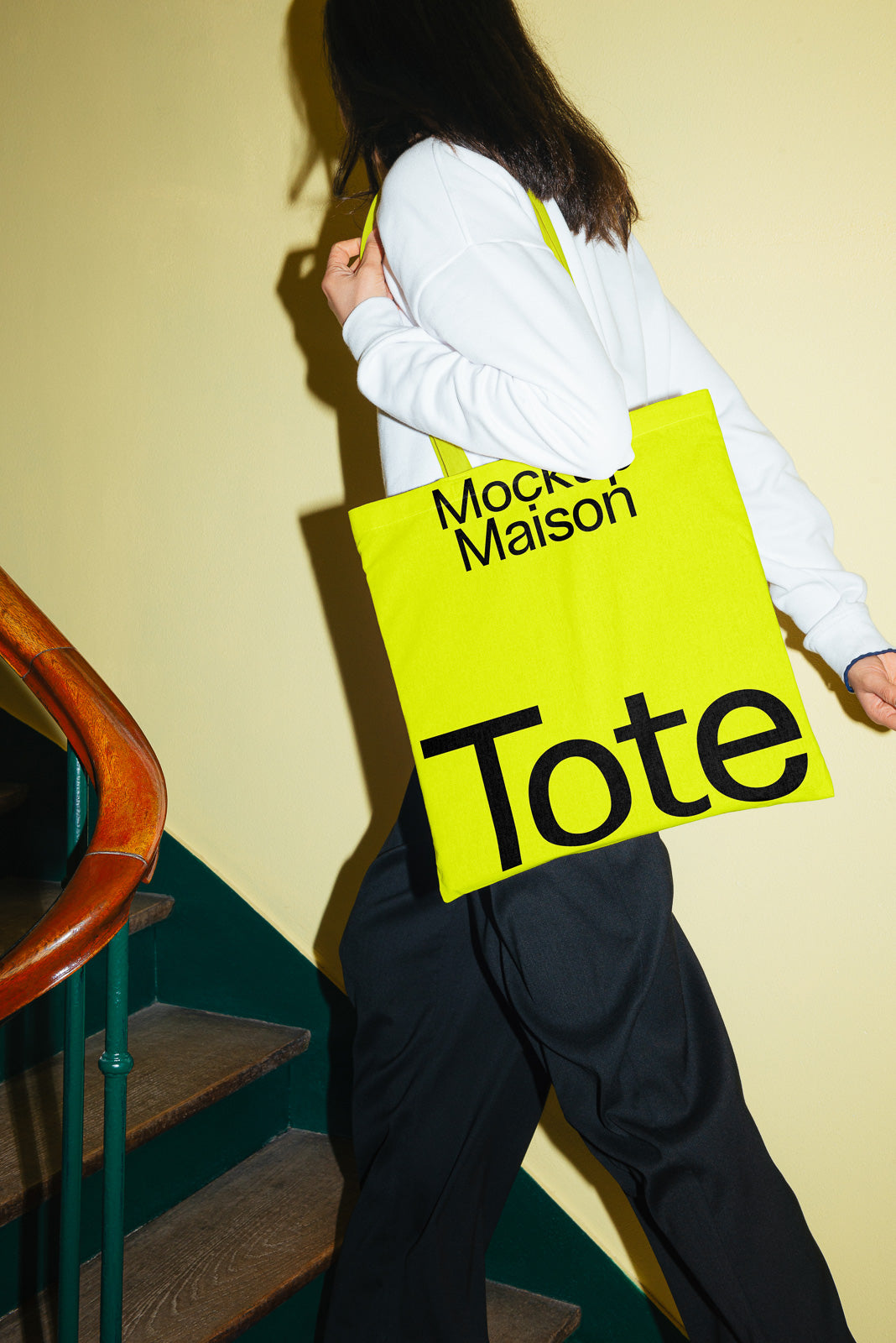 PSD mockup of a person carrying a bright yellow tote bag with "Mockup Maison Tote" text, walking up wooden stairs with a curved railing.