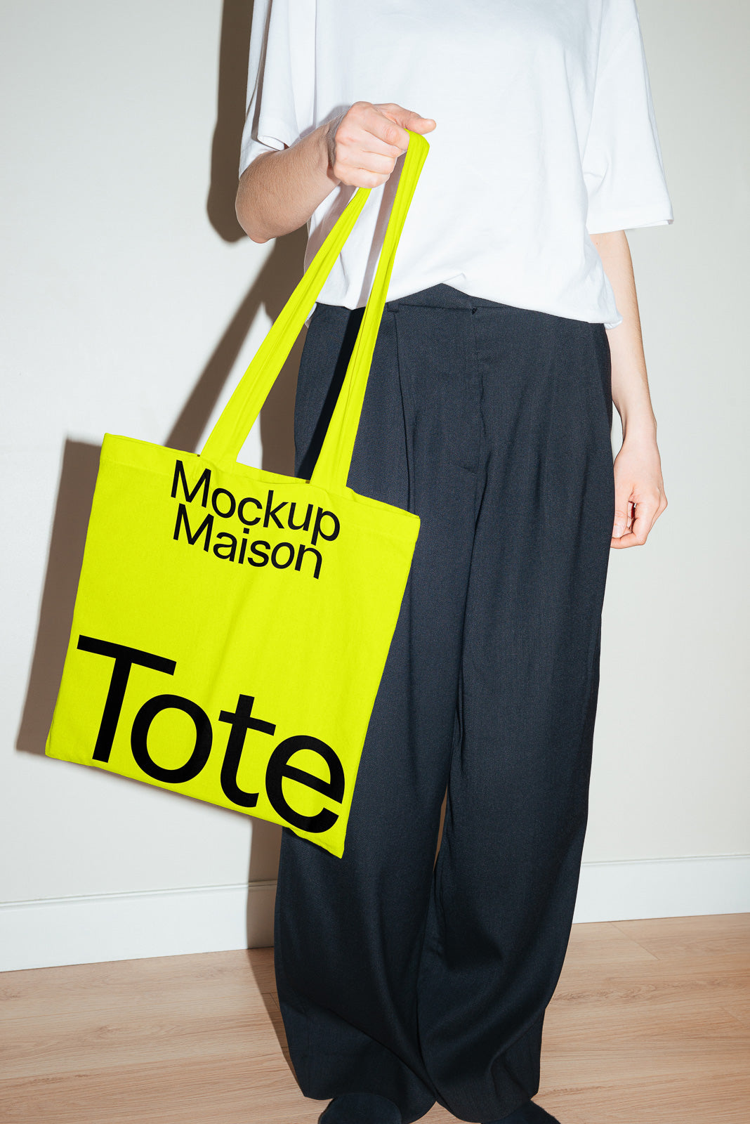 PSD mockup of a person holding a bright yellow tote bag with "Mockup Maison" text. The person wears a white shirt and dark pants.