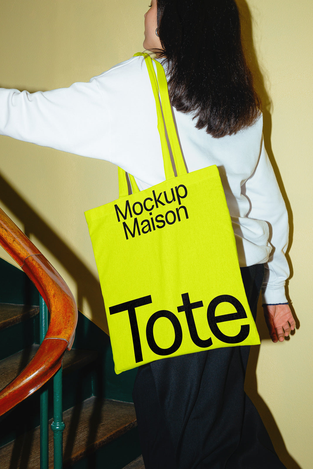 PSD mockup of a person holding a bright yellow tote bag with bold black text, ascending a staircase with a wooden railing.