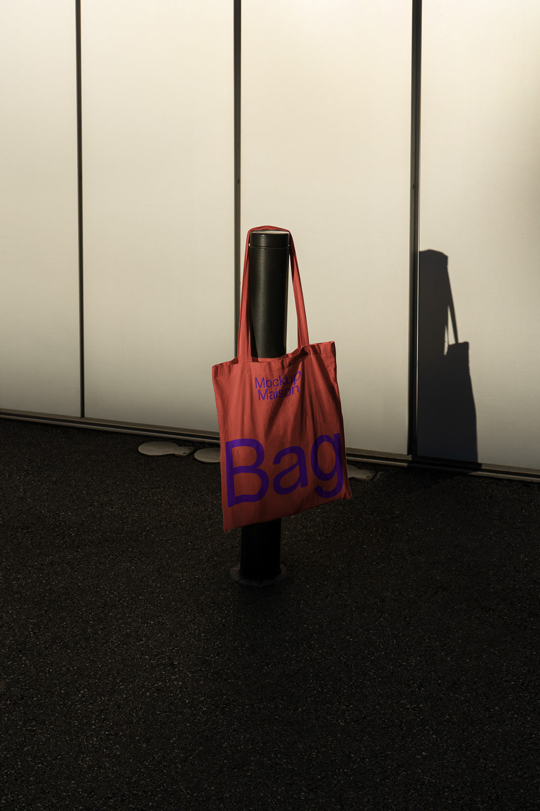 PSD mockup of a red tote bag hanging on a black bollard, casting a shadow on the pavement against a light backdrop.