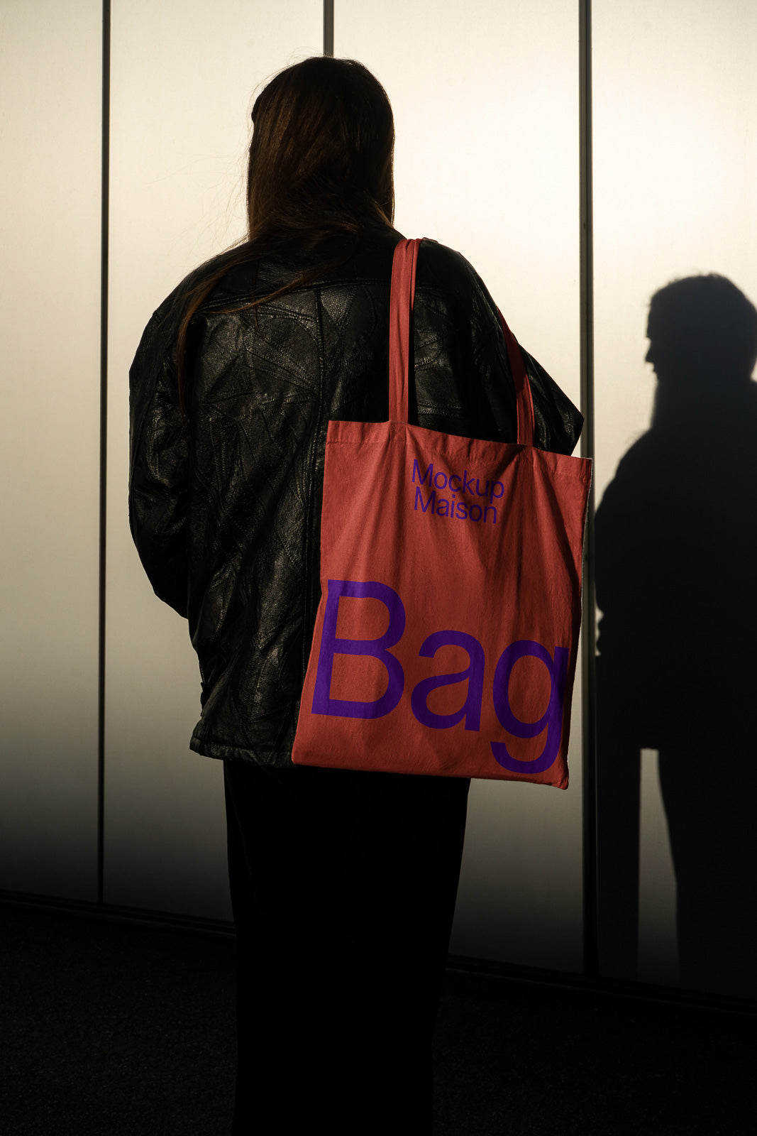 PSD mockup of a person wearing a dark jacket, holding a pink tote bag with bold text "Mockup Maison Bag" on it, against a wall casting a shadow.