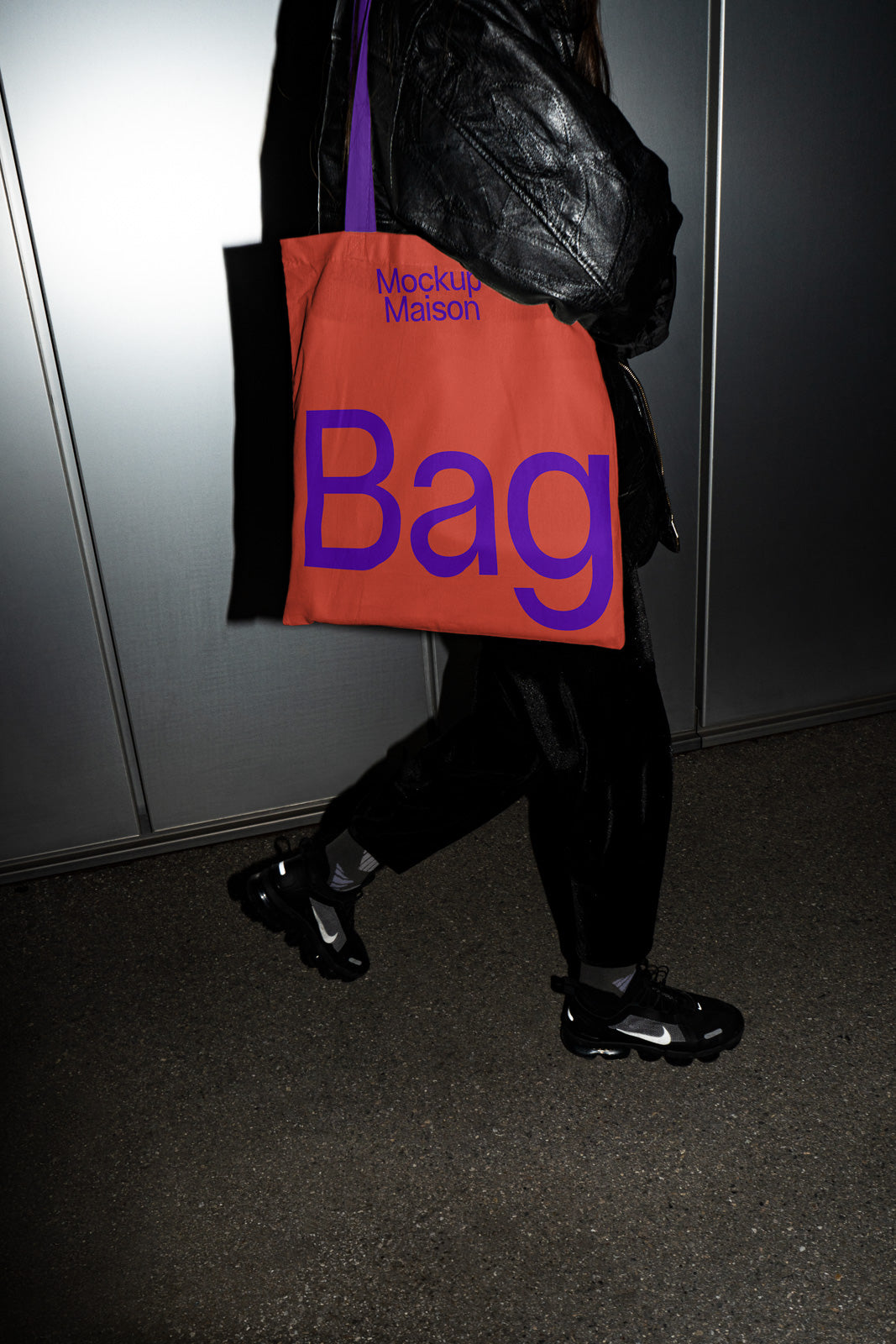 PSD mockup of a person in a black leather jacket holding an orange tote bag with purple text 'Mockup Maison Bag' against a metallic background.