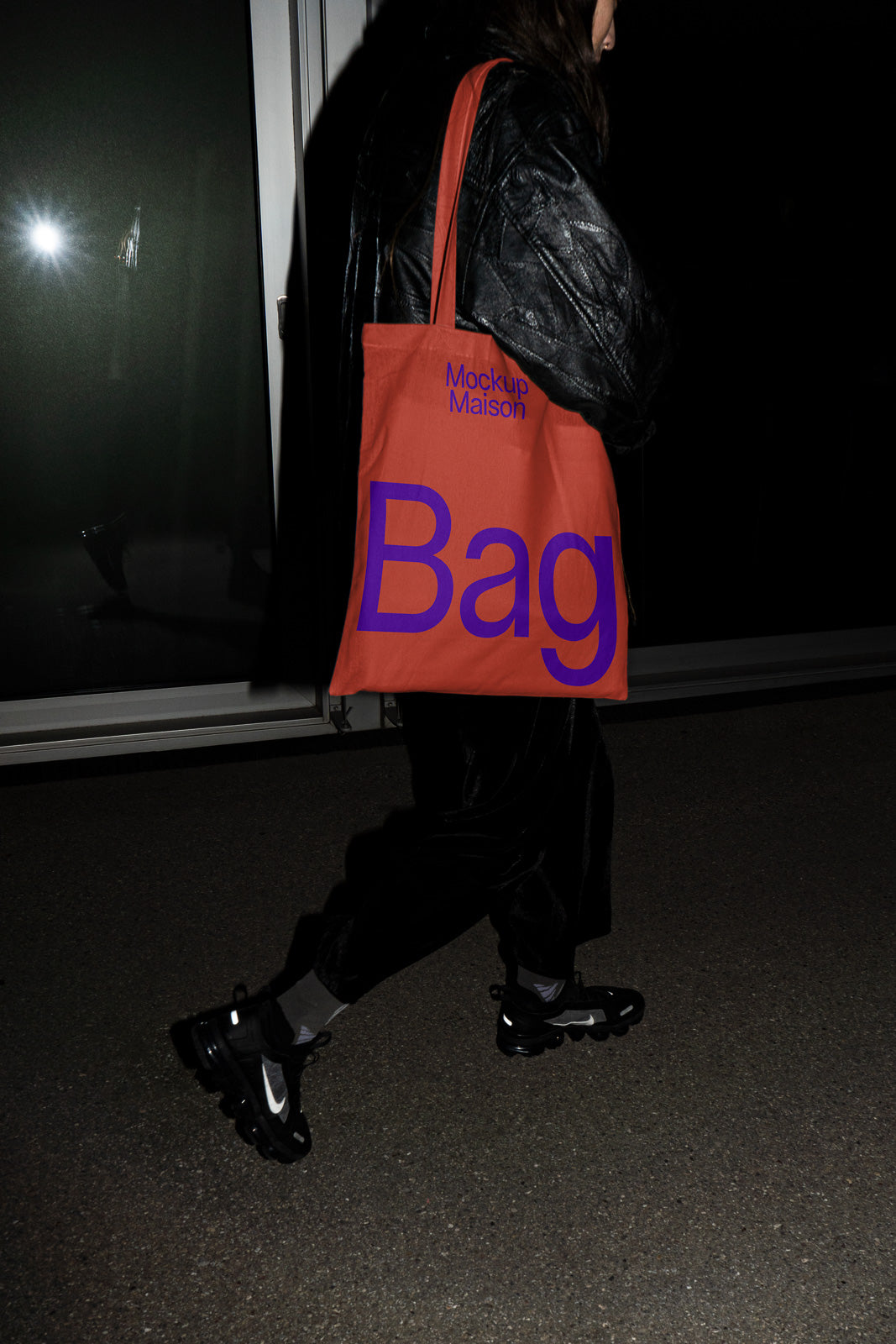 PSD mockup of a red tote bag with "Mockup Maison" and large "Bag" text, carried by a person wearing black leather and sneakers, in nighttime setting.