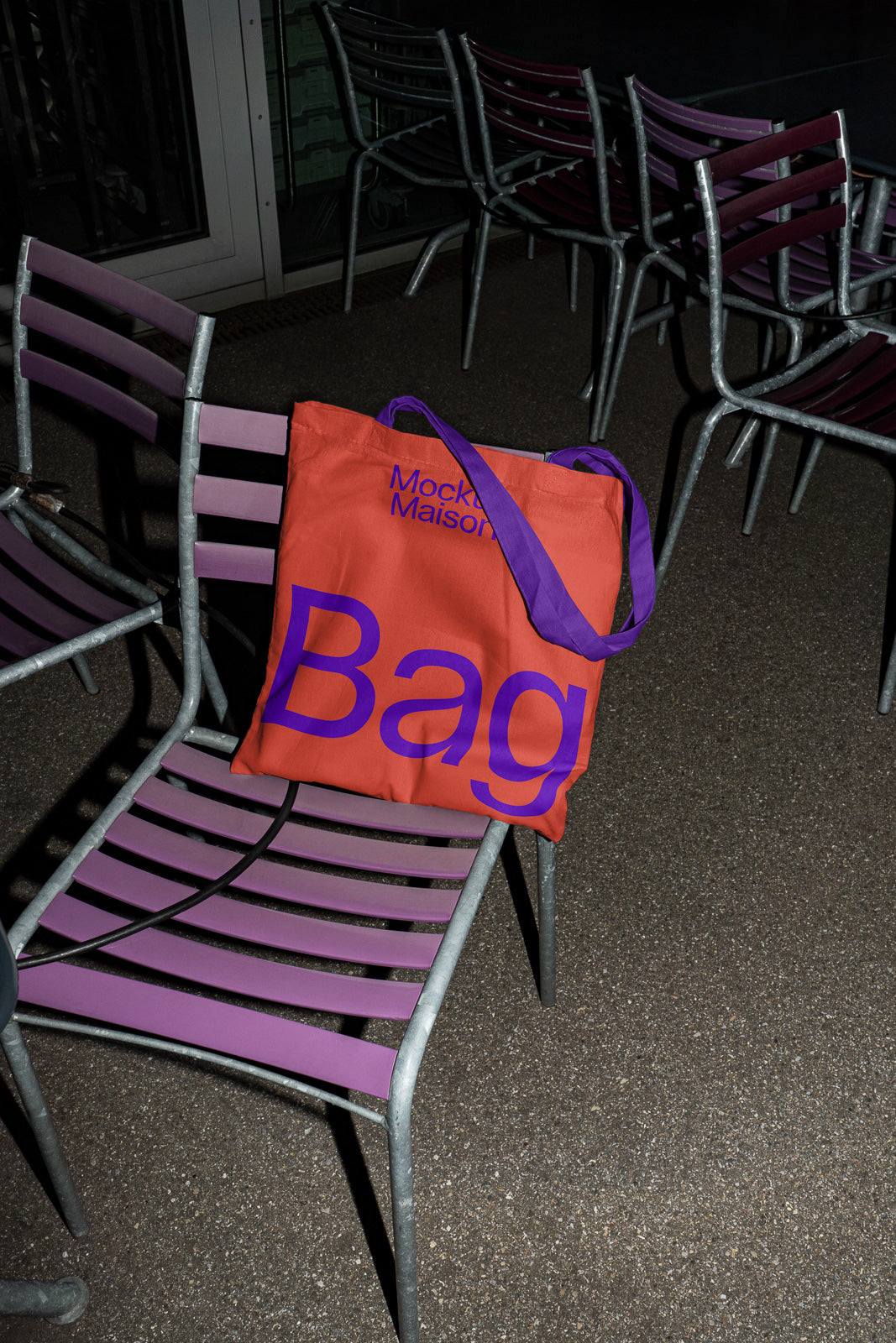 PSD mockup of an orange tote bag with purple text on metal and purple-striped chairs in an outdoor setting at night.