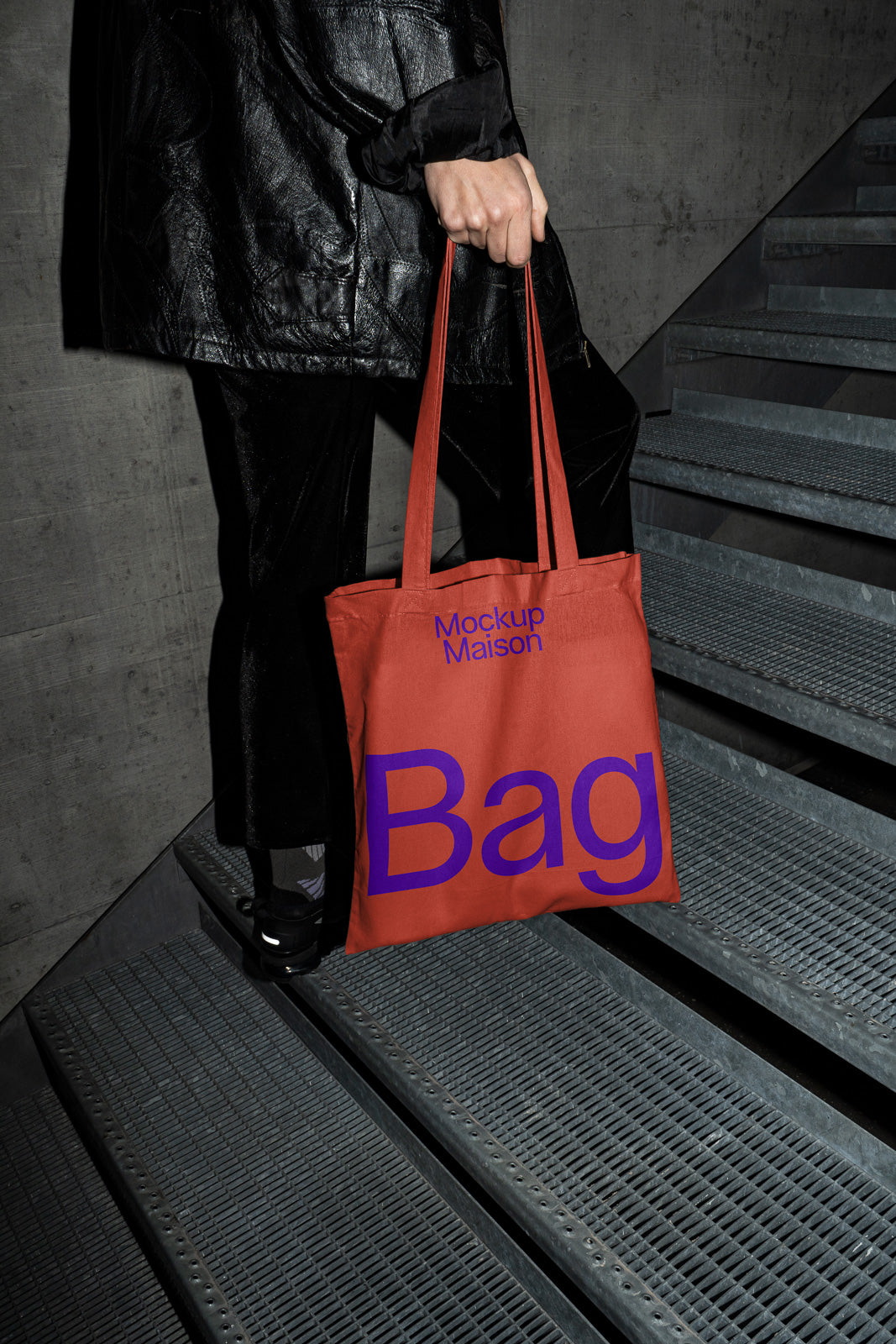 PSD mockup of a person holding an orange tote bag with purple text, walking up metal stairs in a dimly lit industrial setting.