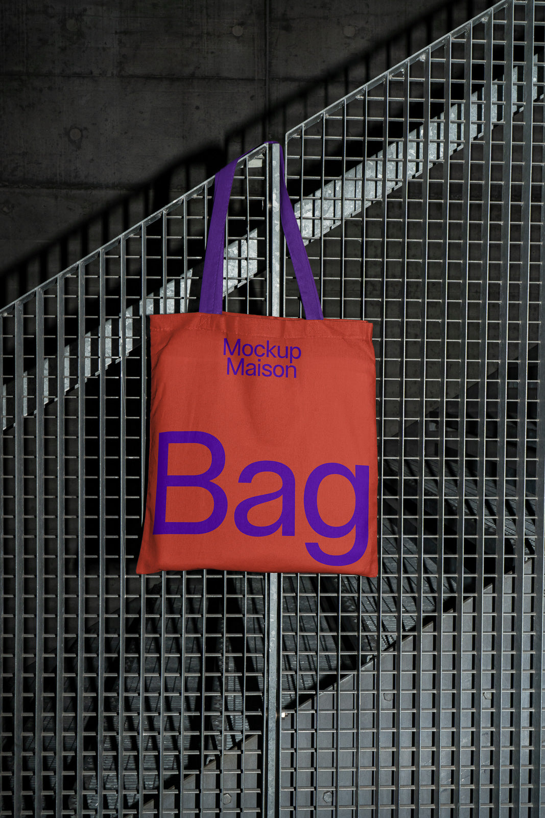 PSD mockup of a red tote bag with purple handles, featuring bold purple text that says "Mockup Maison Bag," hanging on a metal fence background.